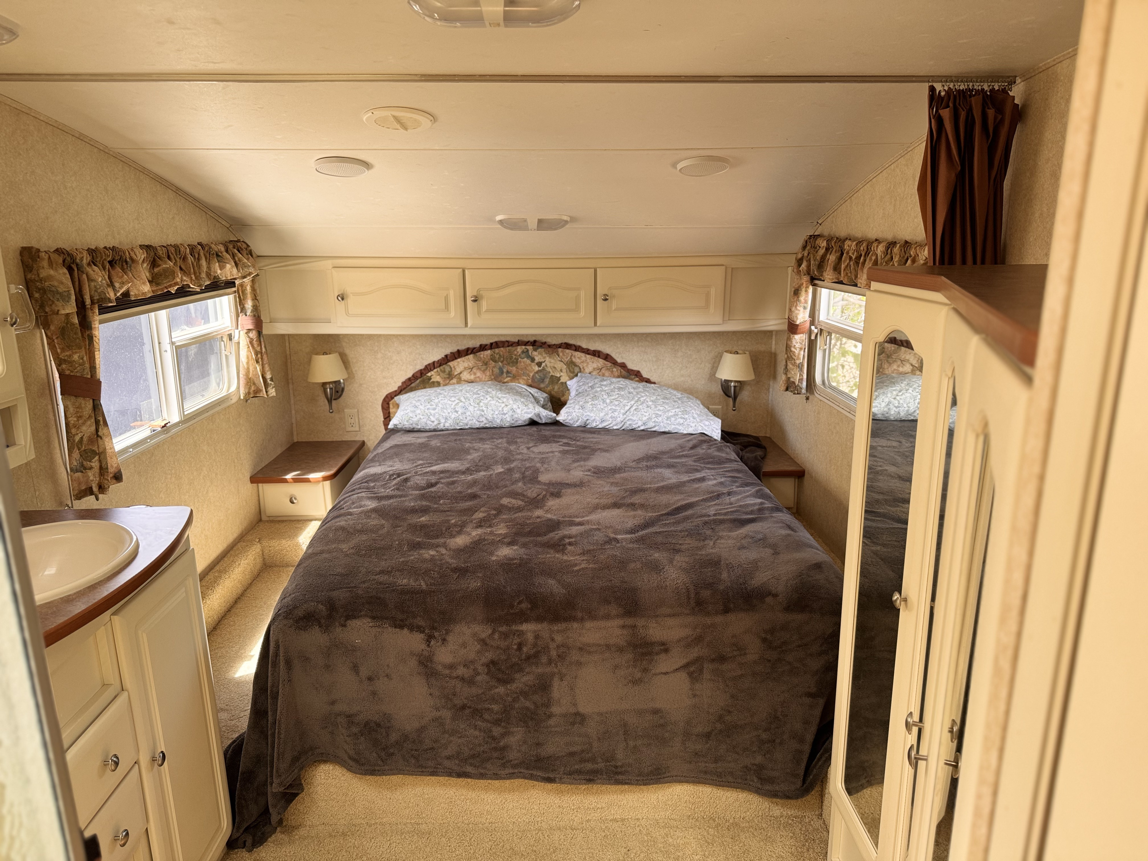 Spacious RV rental bedroom with a brown blanket on the bed, overhead storage, and a sink on the left.. Keystone RV Outback Sydney 2006