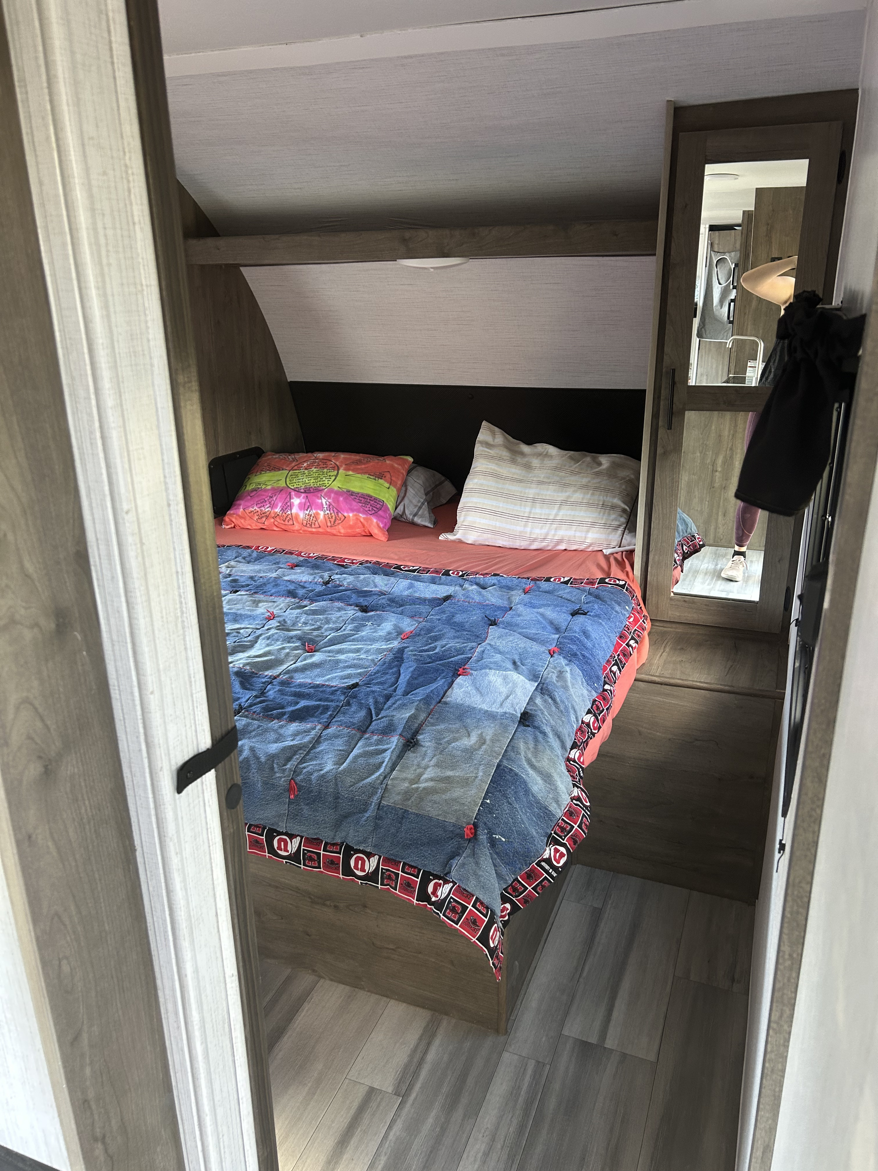 RV rental bedroom with a unique denim quilt, colorful pillows, and a mirror reflecting the adjacent kitchen.. Heartland RVs Pioneer 2022