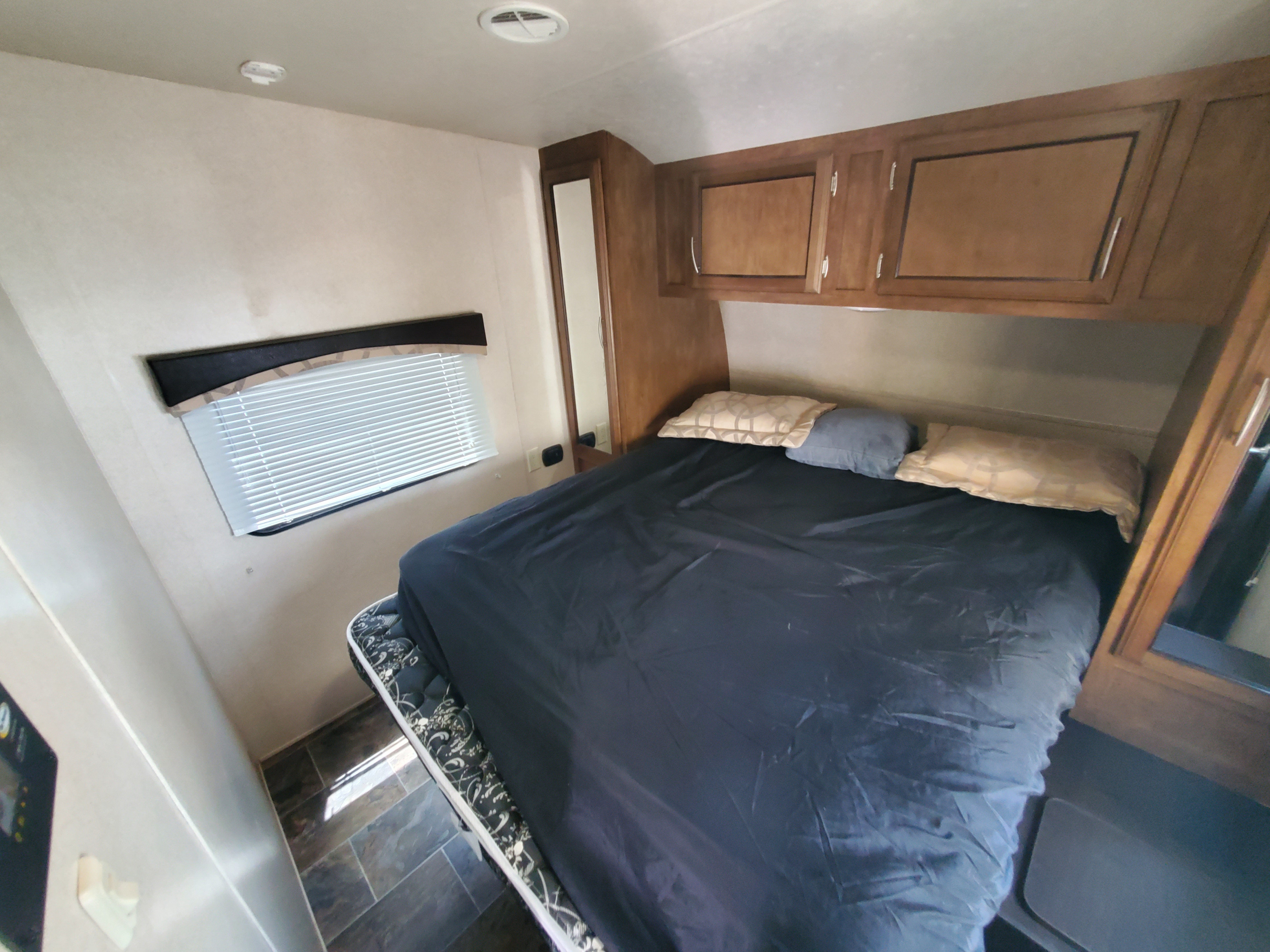 RV rental bedroom features a bed with pillows, wood overhead cabinets, a window with blinds, and a mirror.. Forest River shockwave 2018