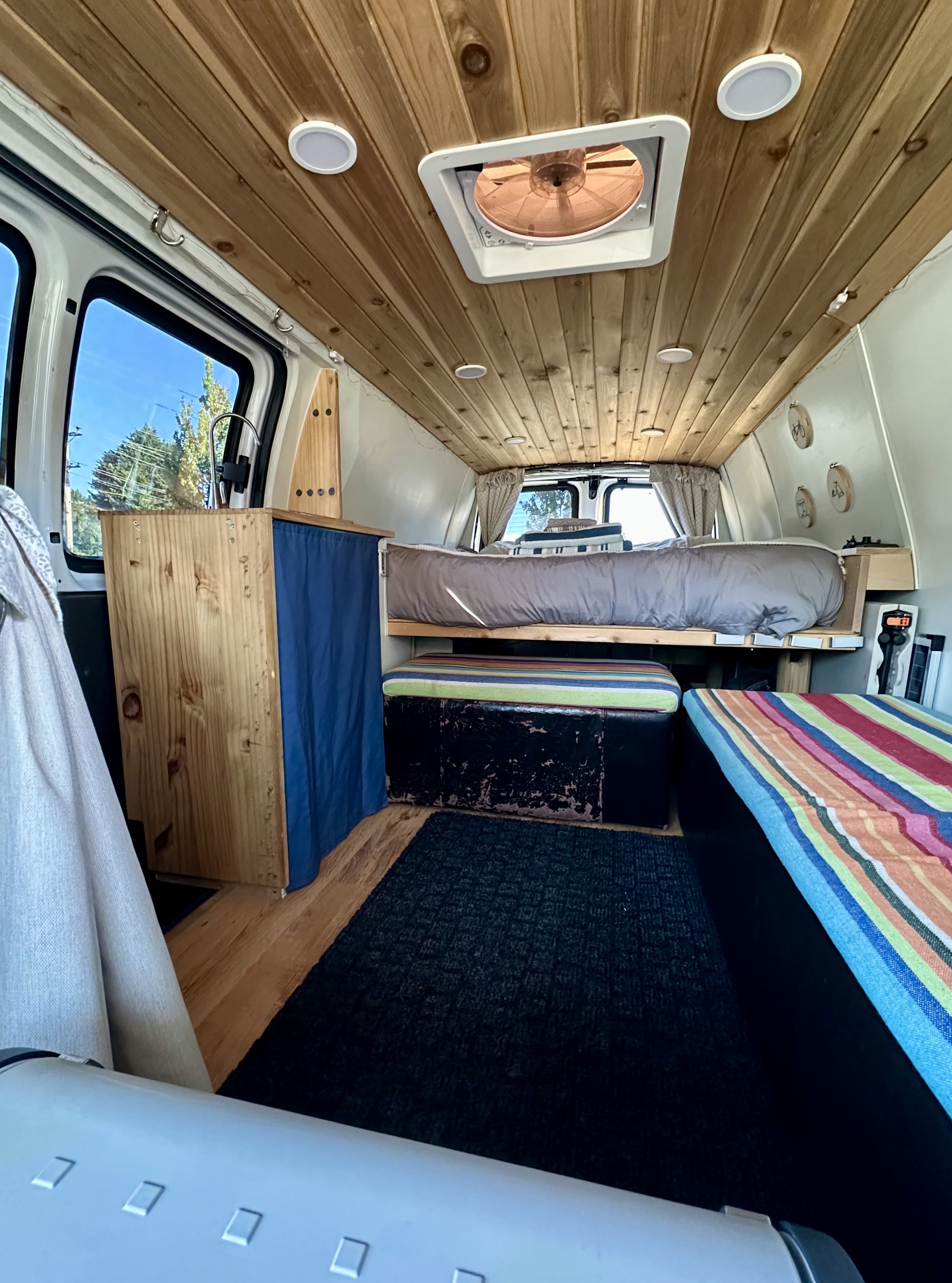 This RV rental shows a cozy camper van interior. Features include a wood plank ceiling, elevated bed, and striped seating.. Chevrolet Express 3500 2006