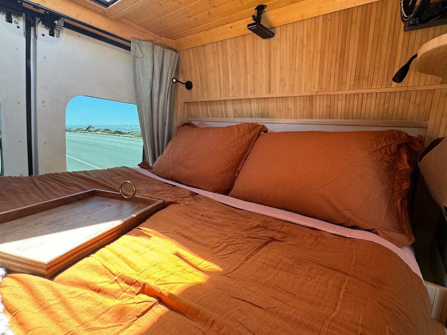 Experience your RV rental adventure! This cozy camper features a comfortable bed with rust linens, wooden walls, and a scenic ocean road view.. Dodge Ram Promaster 2500 2020