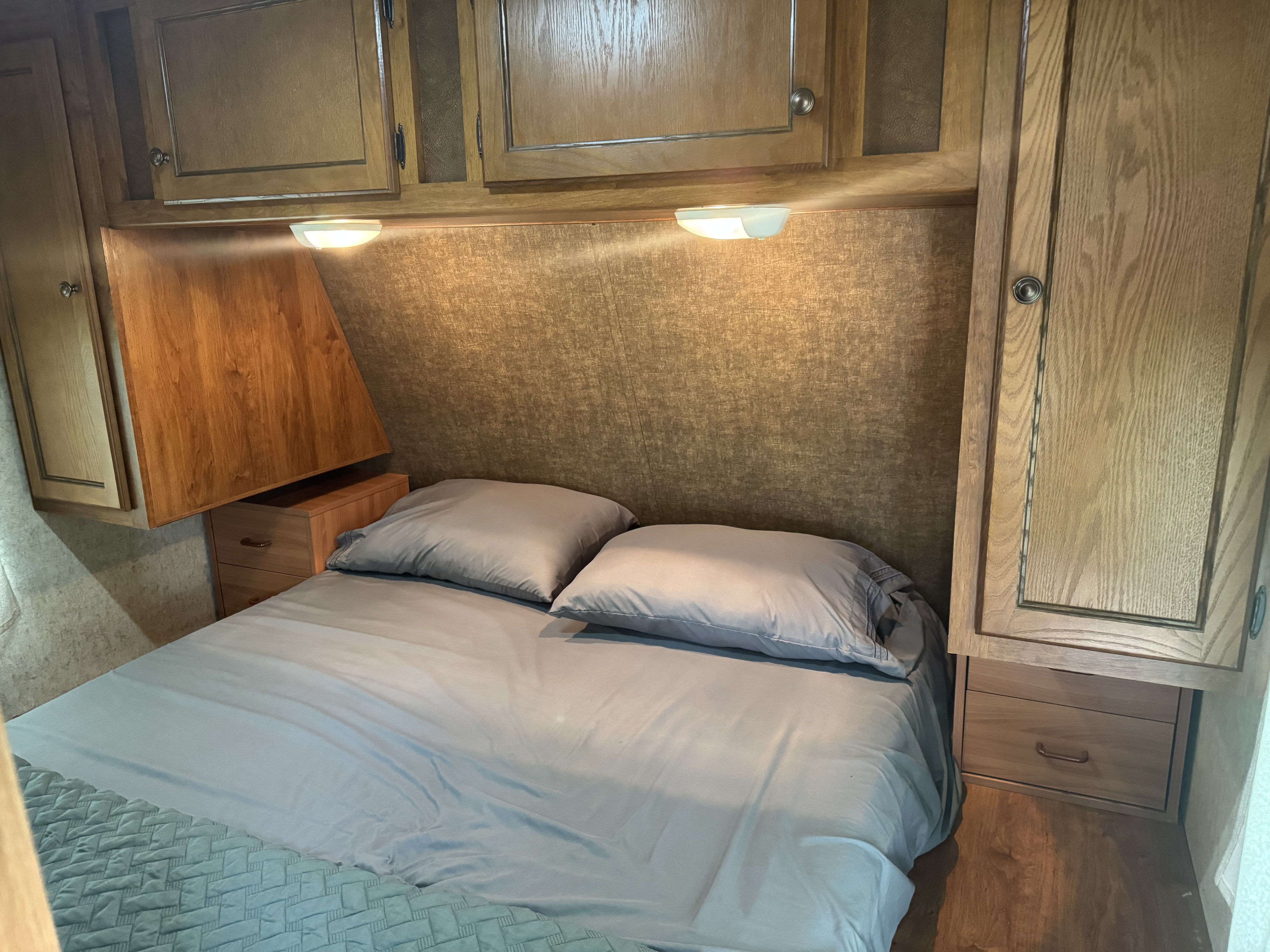 Inside an RV rental, a cozy bedroom with light bedding, wooden overhead cabinets, reading lights, and built-in bedside storage.. Heartland RVs Trail Runner 2014