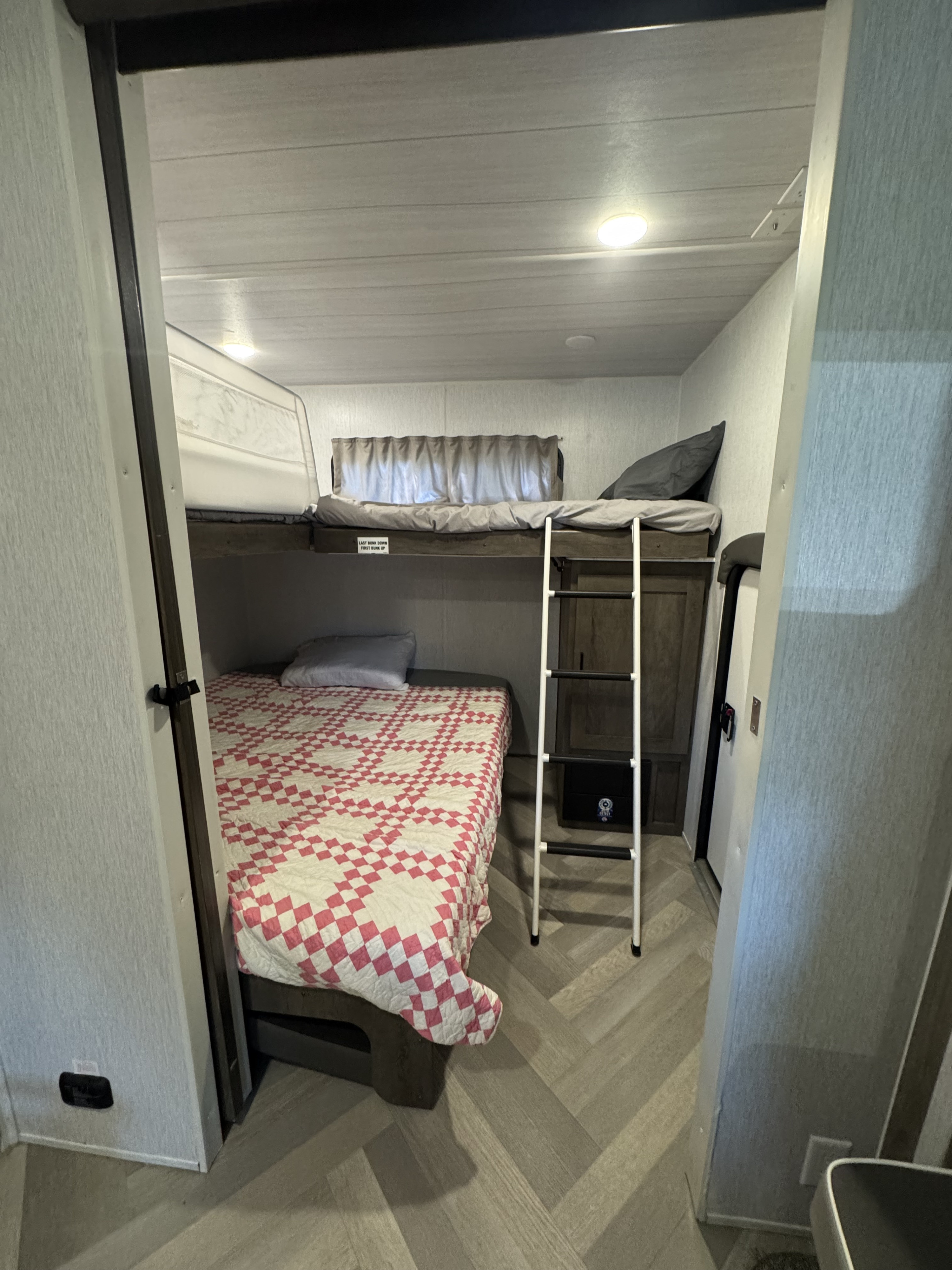 Inside an RV rental: comfortable bunk beds, the lower with a checkered quilt, the upper with a gray comforter, and a ladder.. Forest River Salem 2022