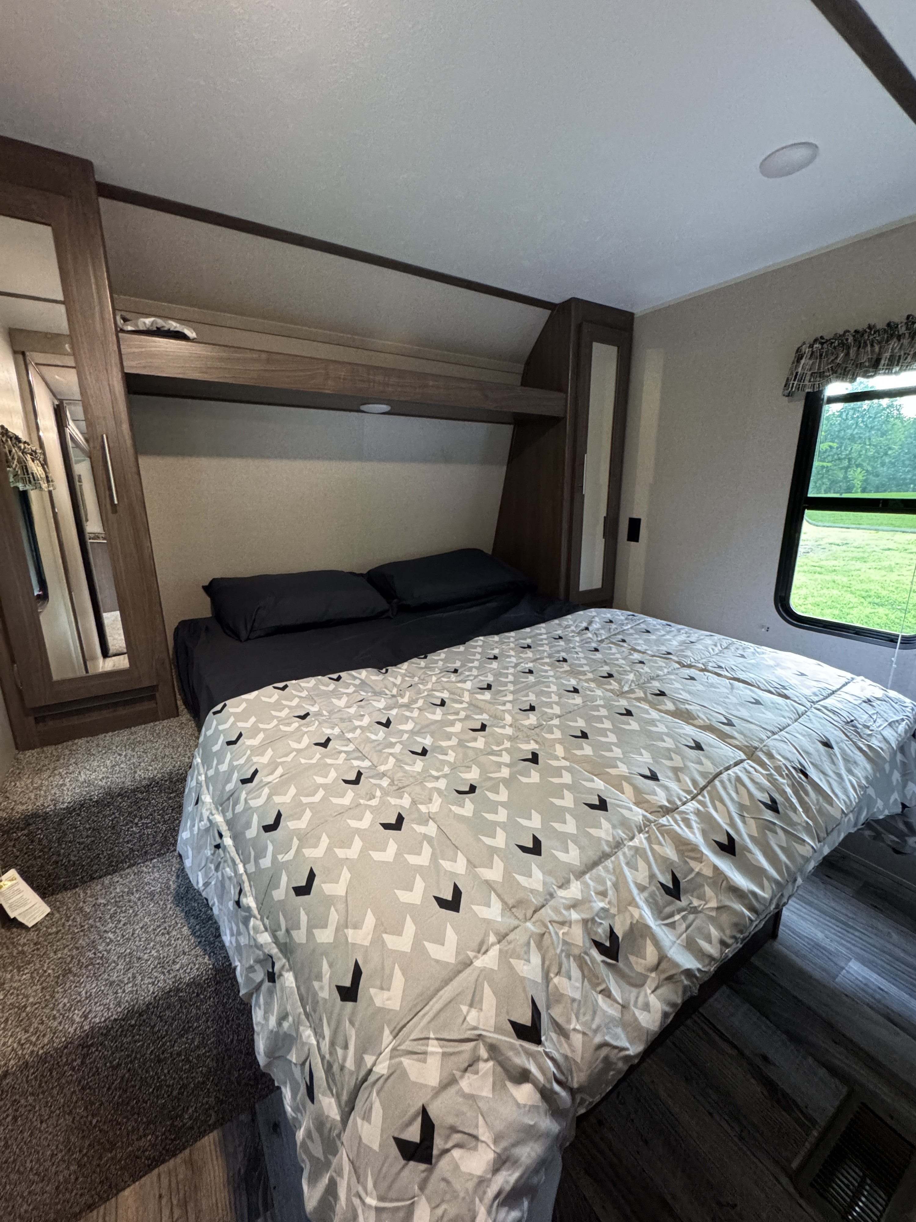 RV rental bedroom with cozy patterned bedding, built-in mirrored cabinets, and a window overlooking lush greenery.. Keystone RV Hideout 2019