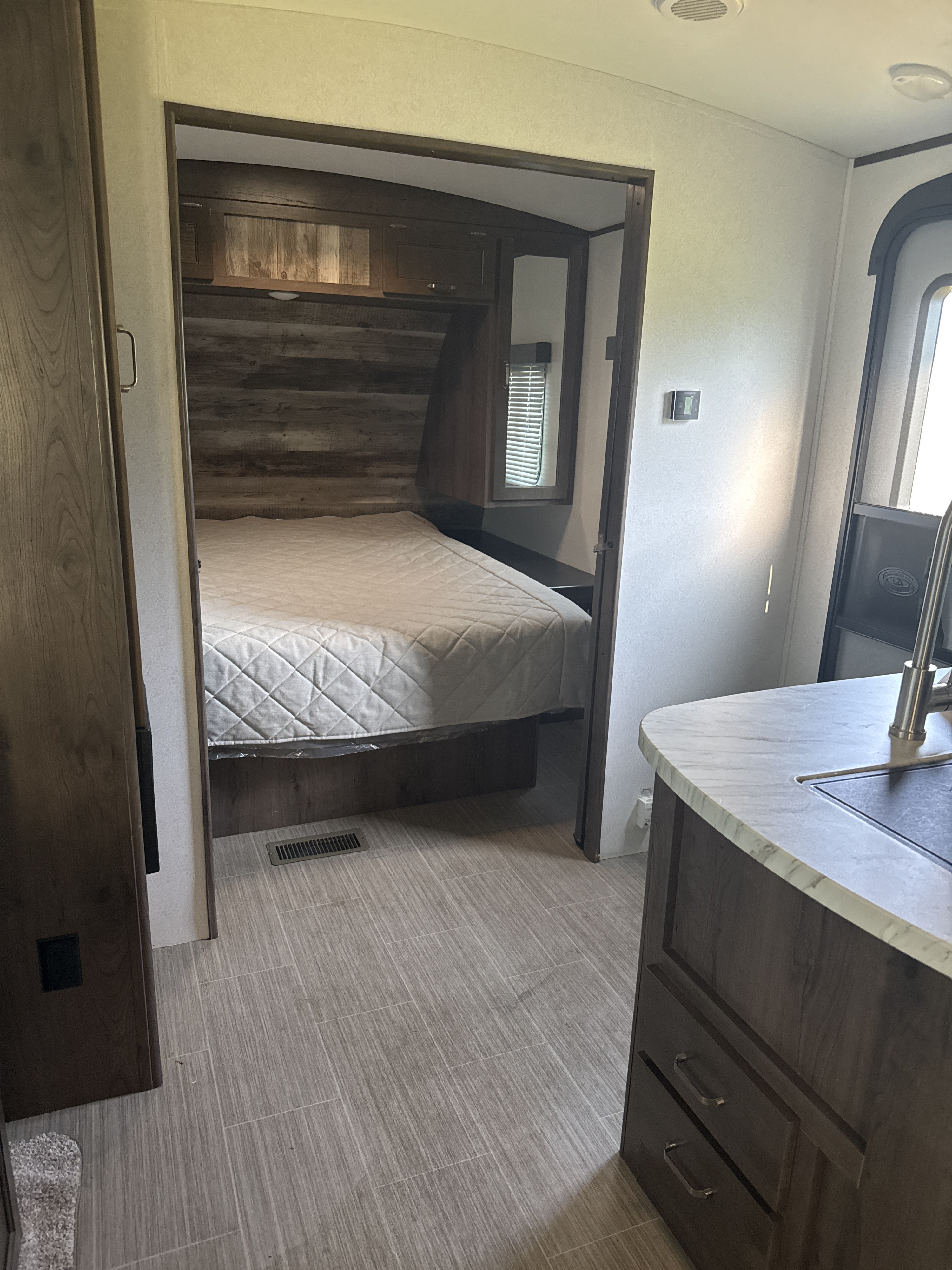 Spacious RV rental interior with a cozy bedroom and kitchen counter. Features wood-look flooring and rustic accents, ideal for travel.. Keystone RV Passport Grand Touring 2021