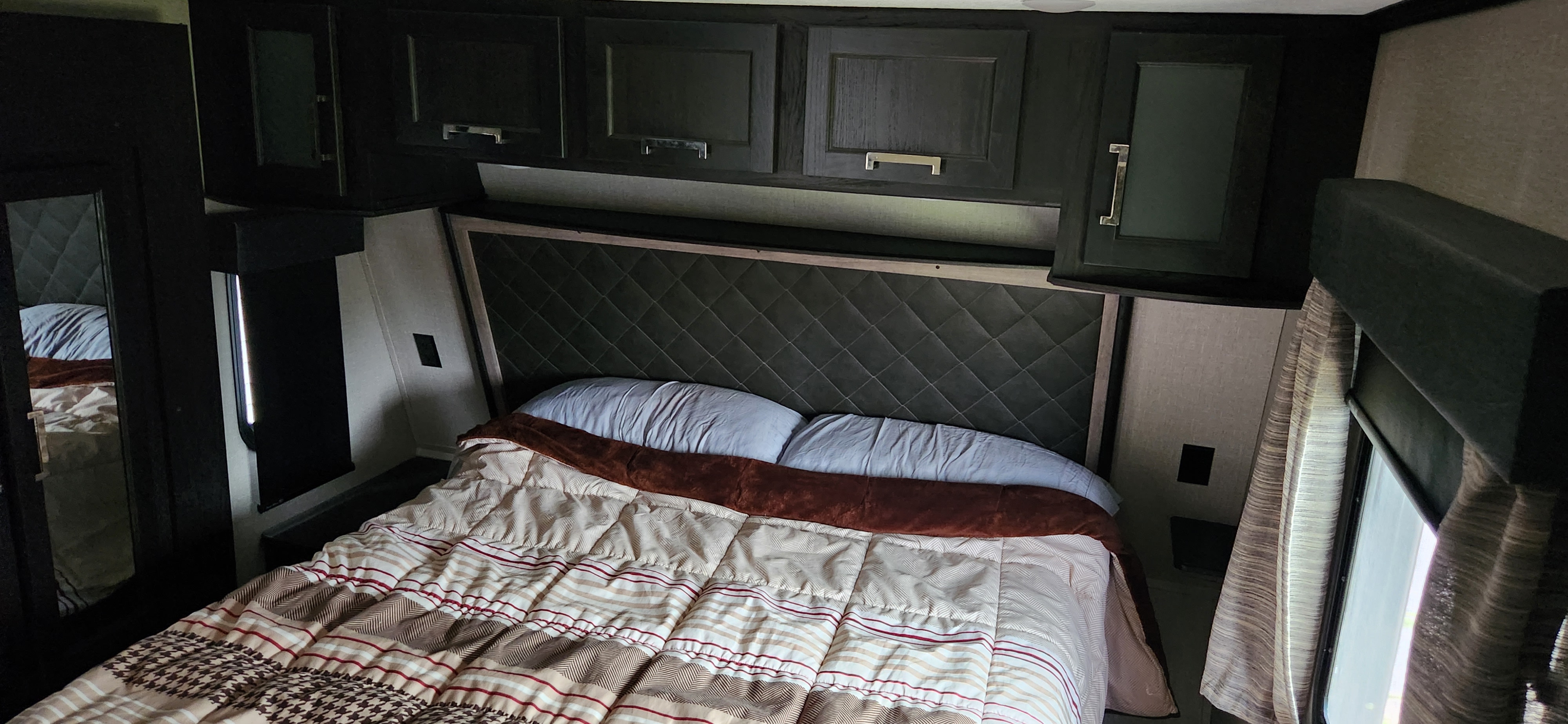 Compact RV rental bedroom showing a bed with patterned comforter, quilted headboard, and overhead cabinets.. Keystone Fuzion 2019