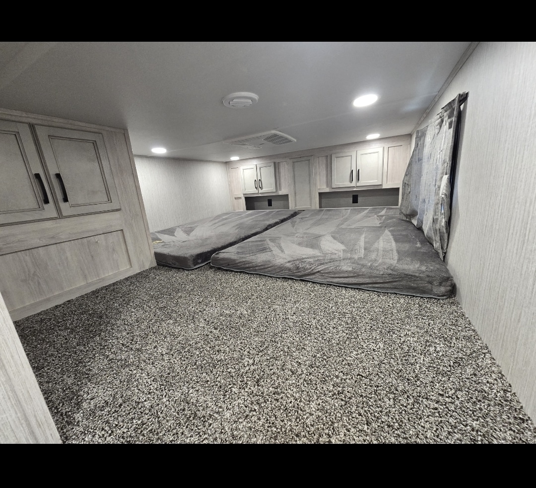 An RV rental's carpeted sleeping loft with two grey beds, light wood cabinets, and overhead lighting.. Forest River Sandpiper 2023