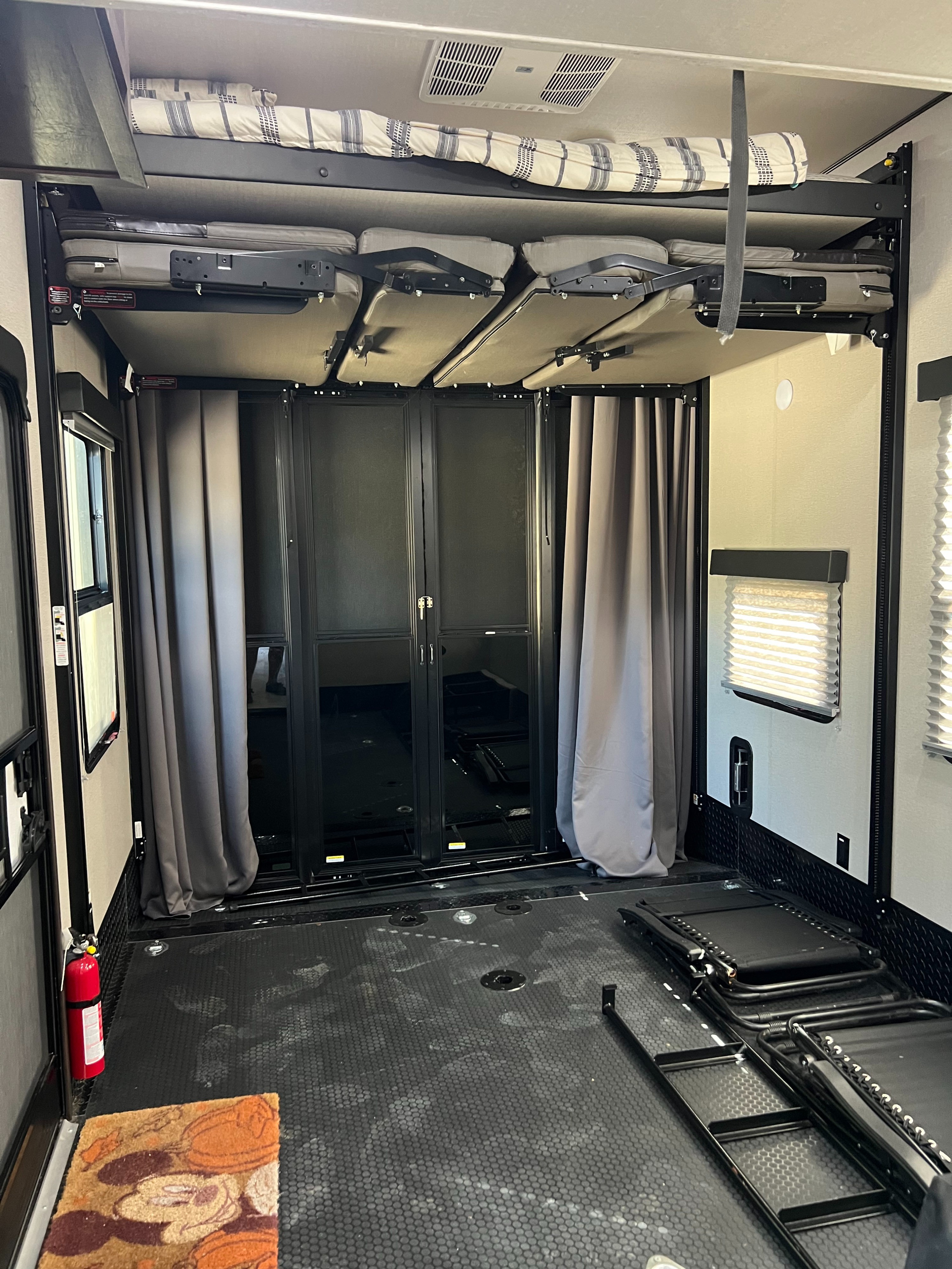 RV rental toy hauler interior with upper bunk, folded lower bunks, and durable cargo floor. Rear screen doors and curtains shown.. Grand Design Momentum 2024