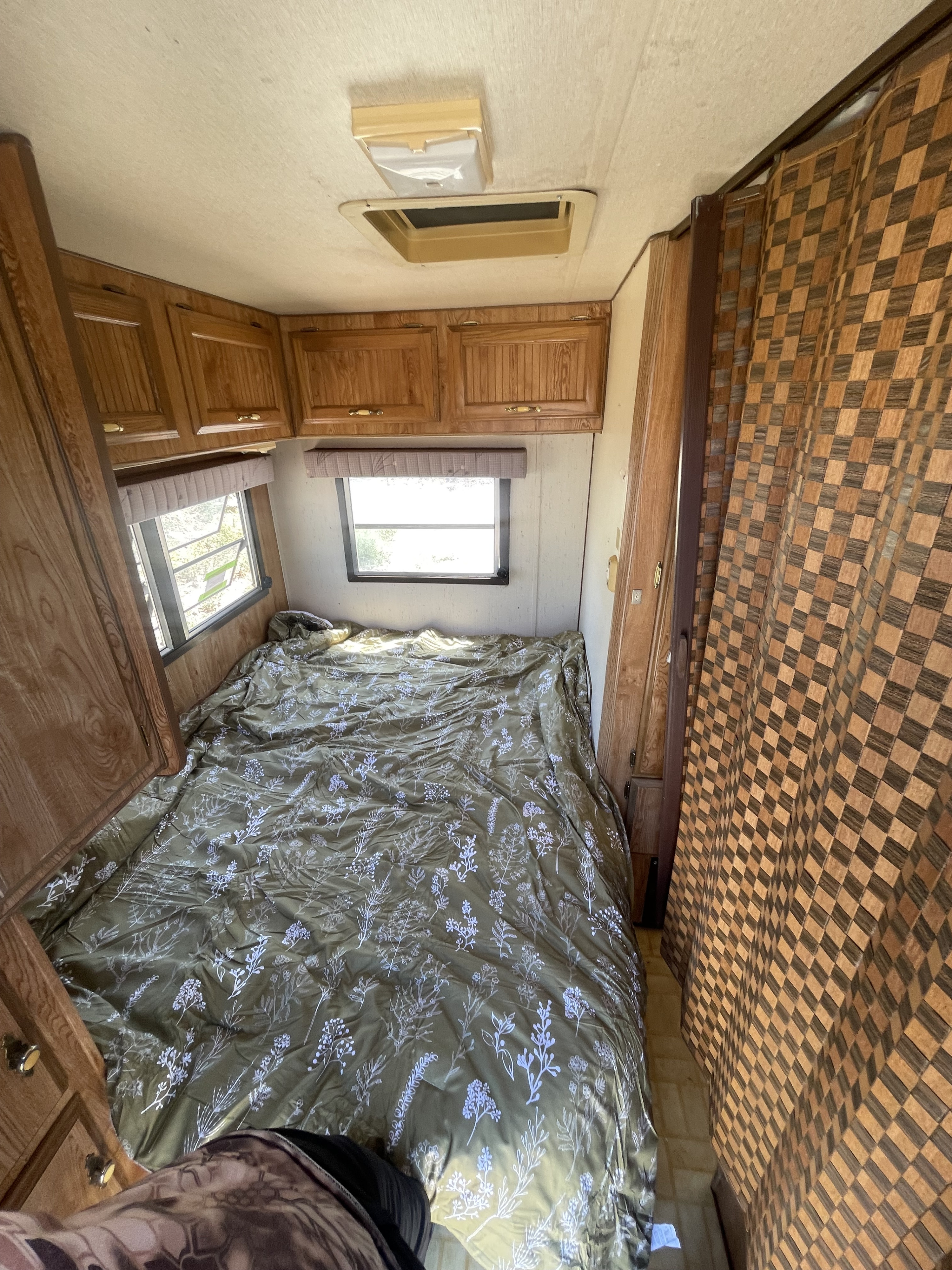 Inside an RV rental: a compact bedroom with a green floral bed, wood cabinets, and checkerboard privacy curtain.. Skyline Aljo 1989