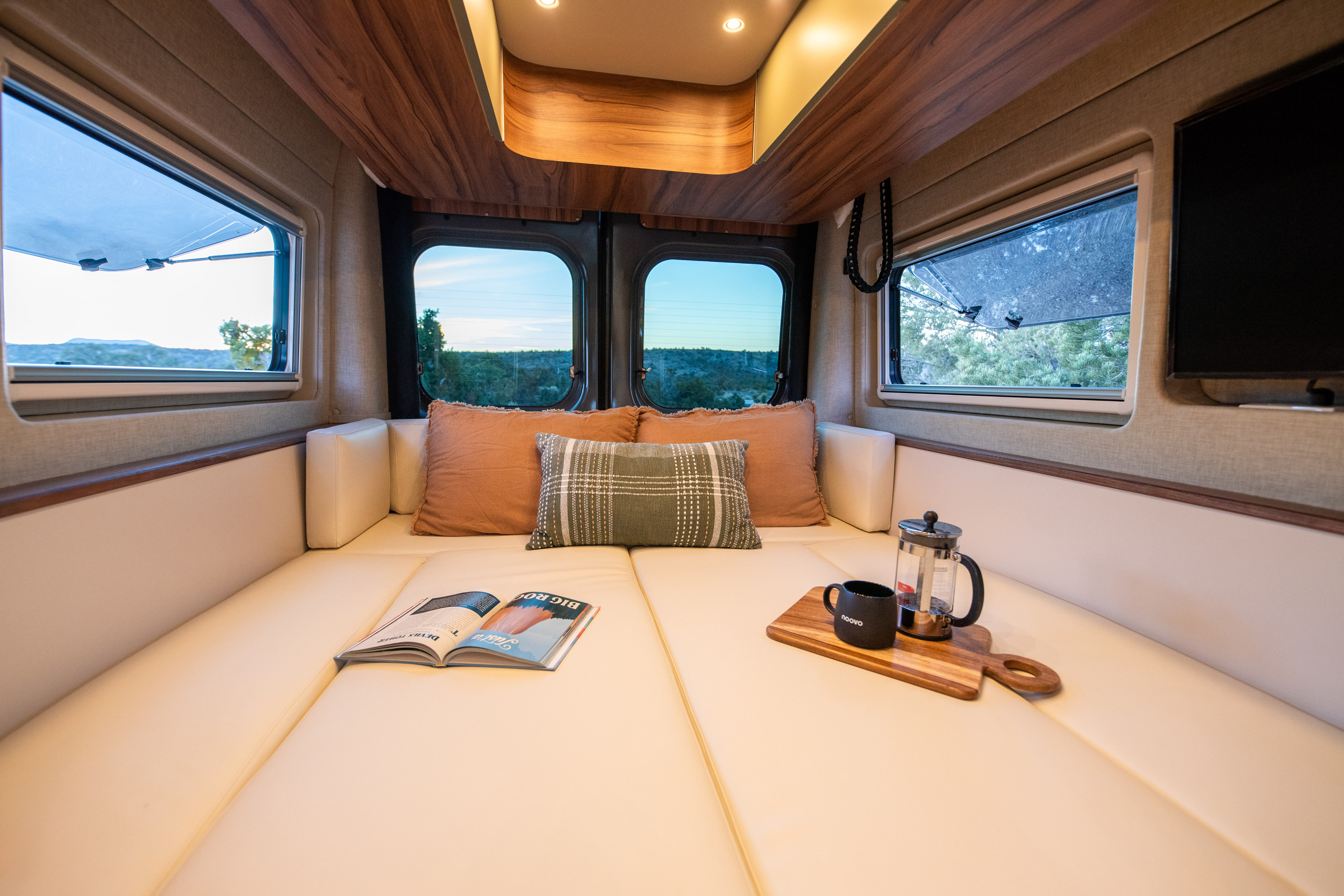 Cozy RV rental interior: lounge bed with coffee, magazine, and scenic views from multiple windows. Perfect for van life adventures!. Noovo Plus LB 2025