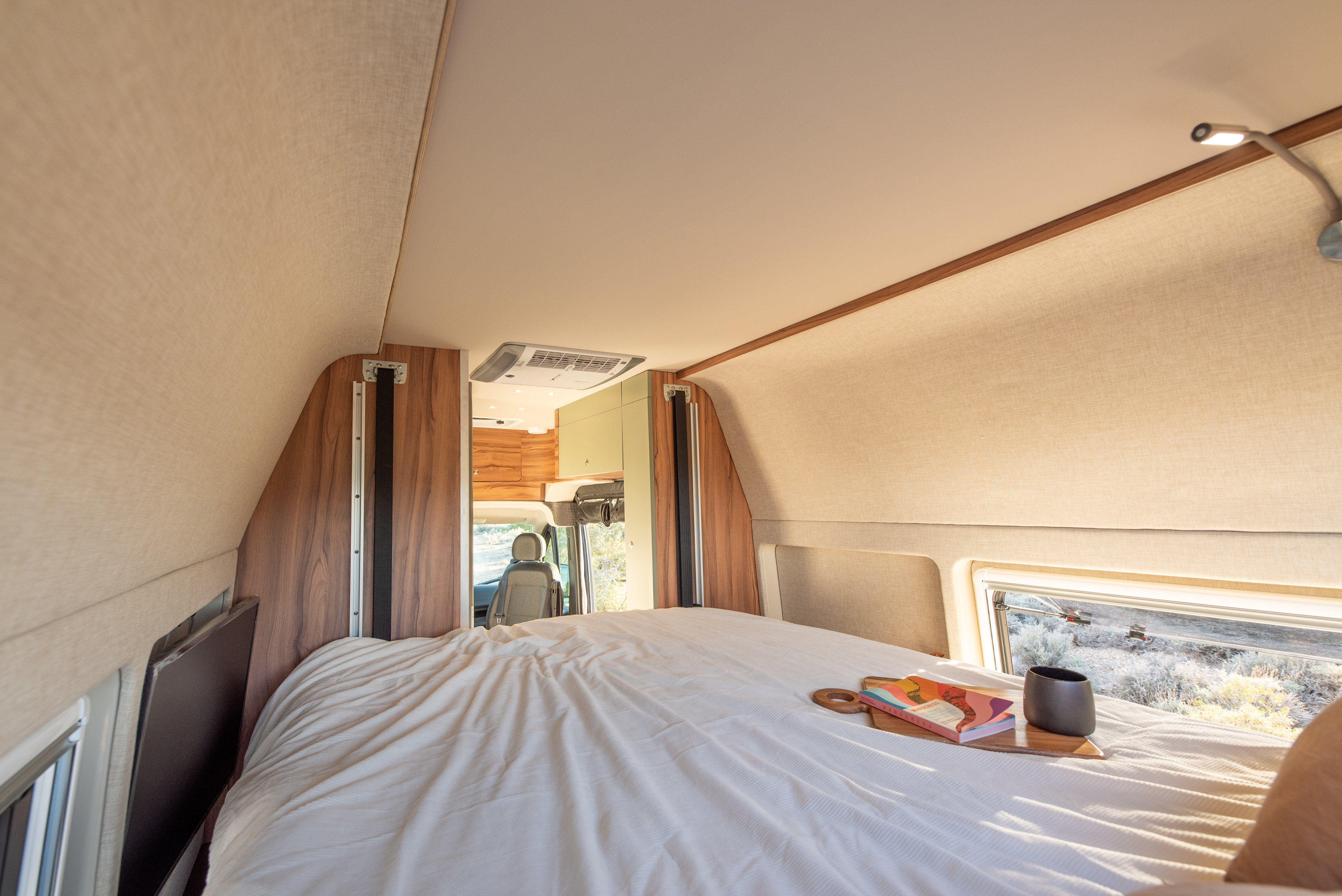 Modern RV rental bedroom: a cozy bed with a book, mug, and a window revealing a scenic outdoor view.. Noovo Plus LB 2025
