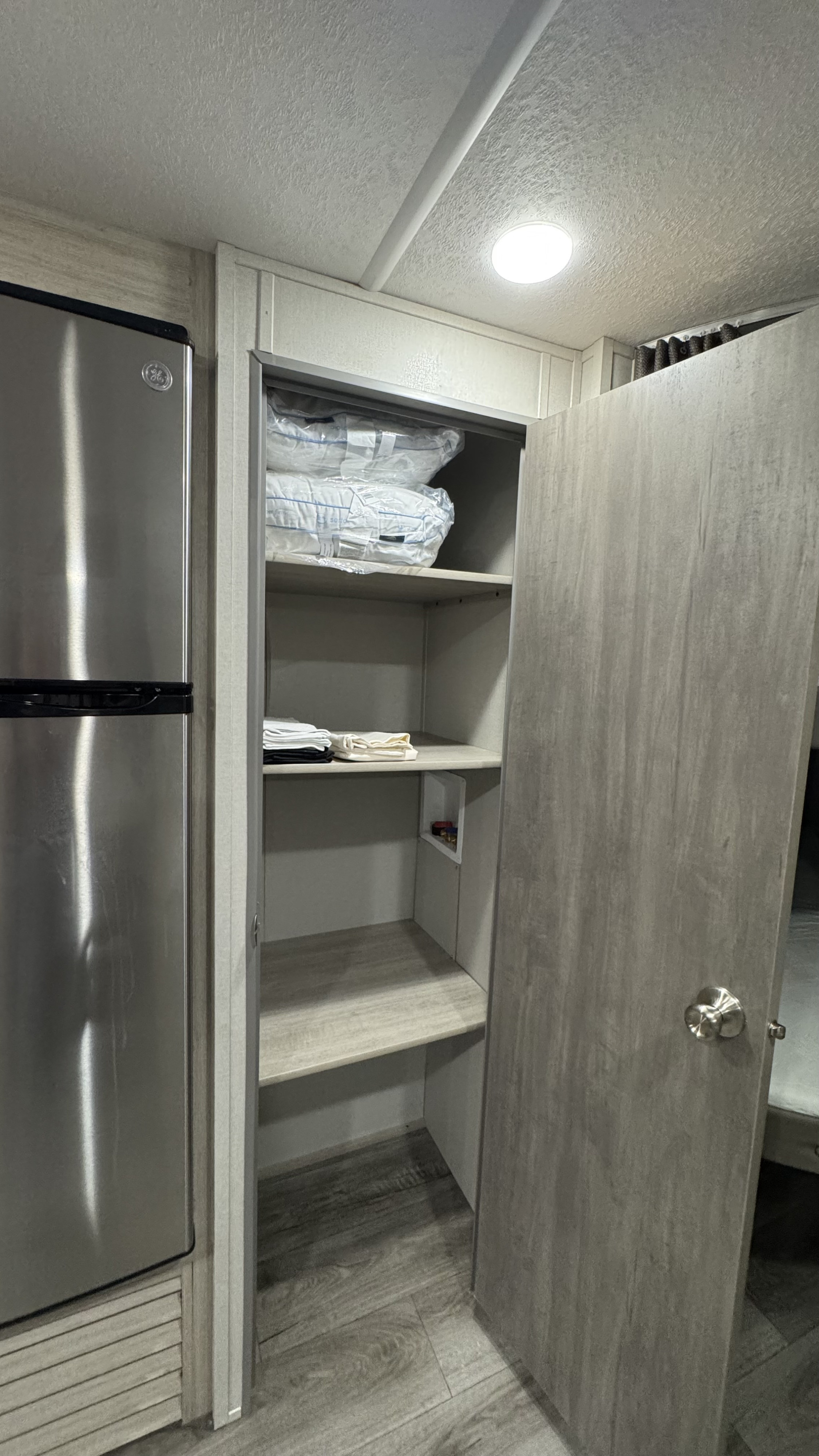 RV rental interior with a GE fridge & open pantry. Shelves hold packaged pillows, folded linens, and plumbing access.. Coachmen Catalina 2025