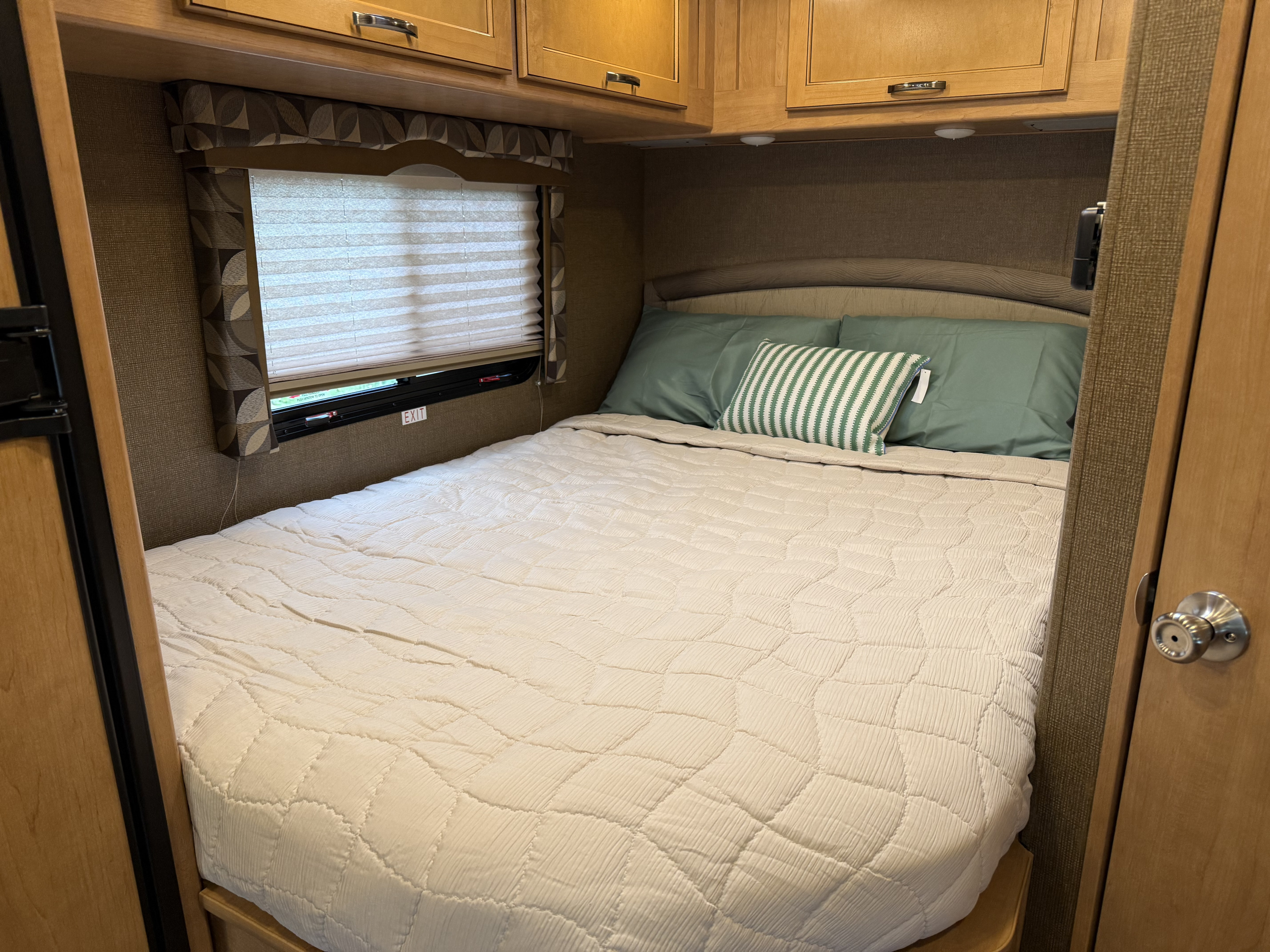 Inside a cozy RV rental bedroom: a bed with soft bedding, overhead cabinets, and a window with blinds.. Thor Chateau 2017