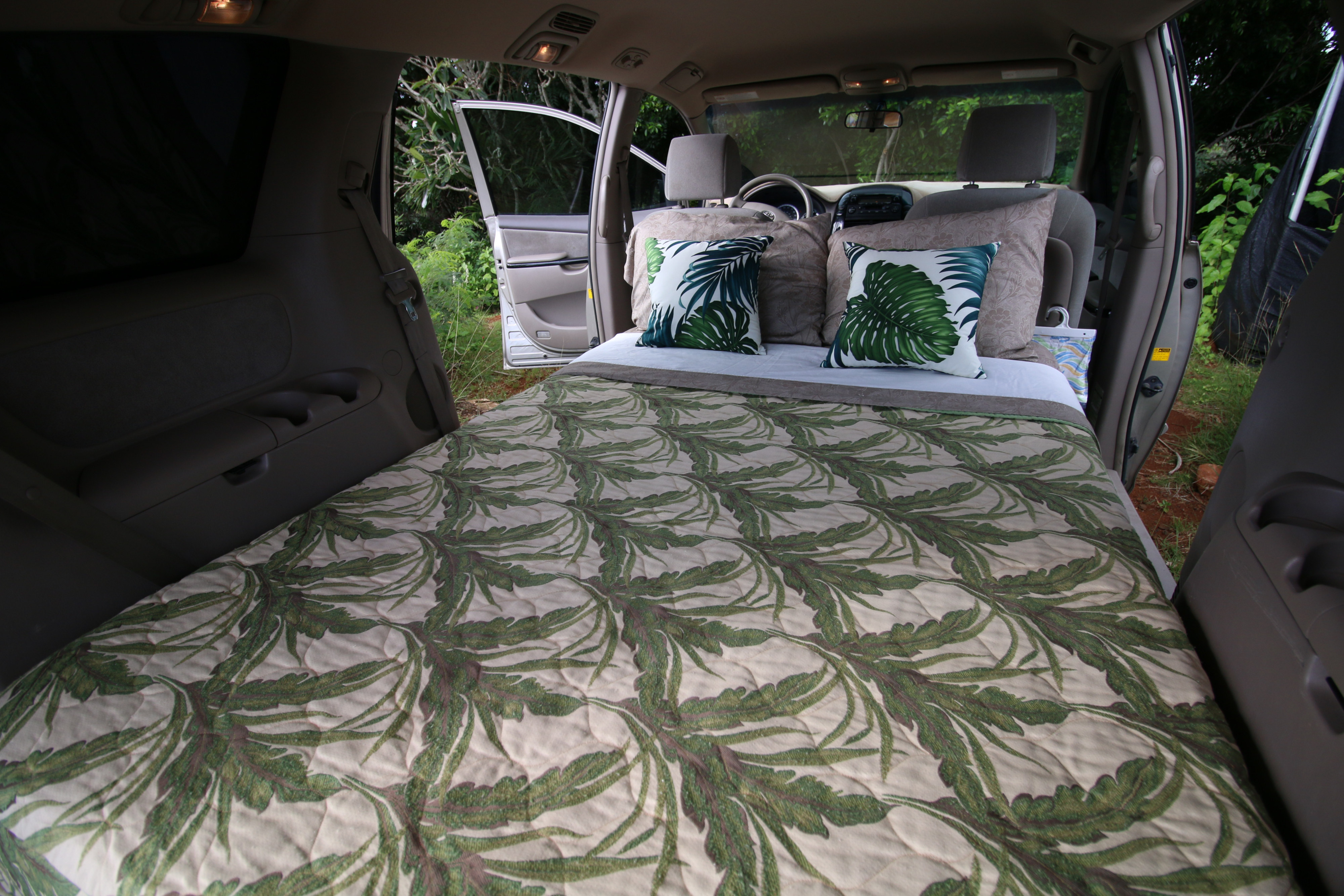 Inside a car converted into a cozy bed with tropical leaf bedding, ready for an outdoor RV rental escape.. Toyota Sienna Camper 2004