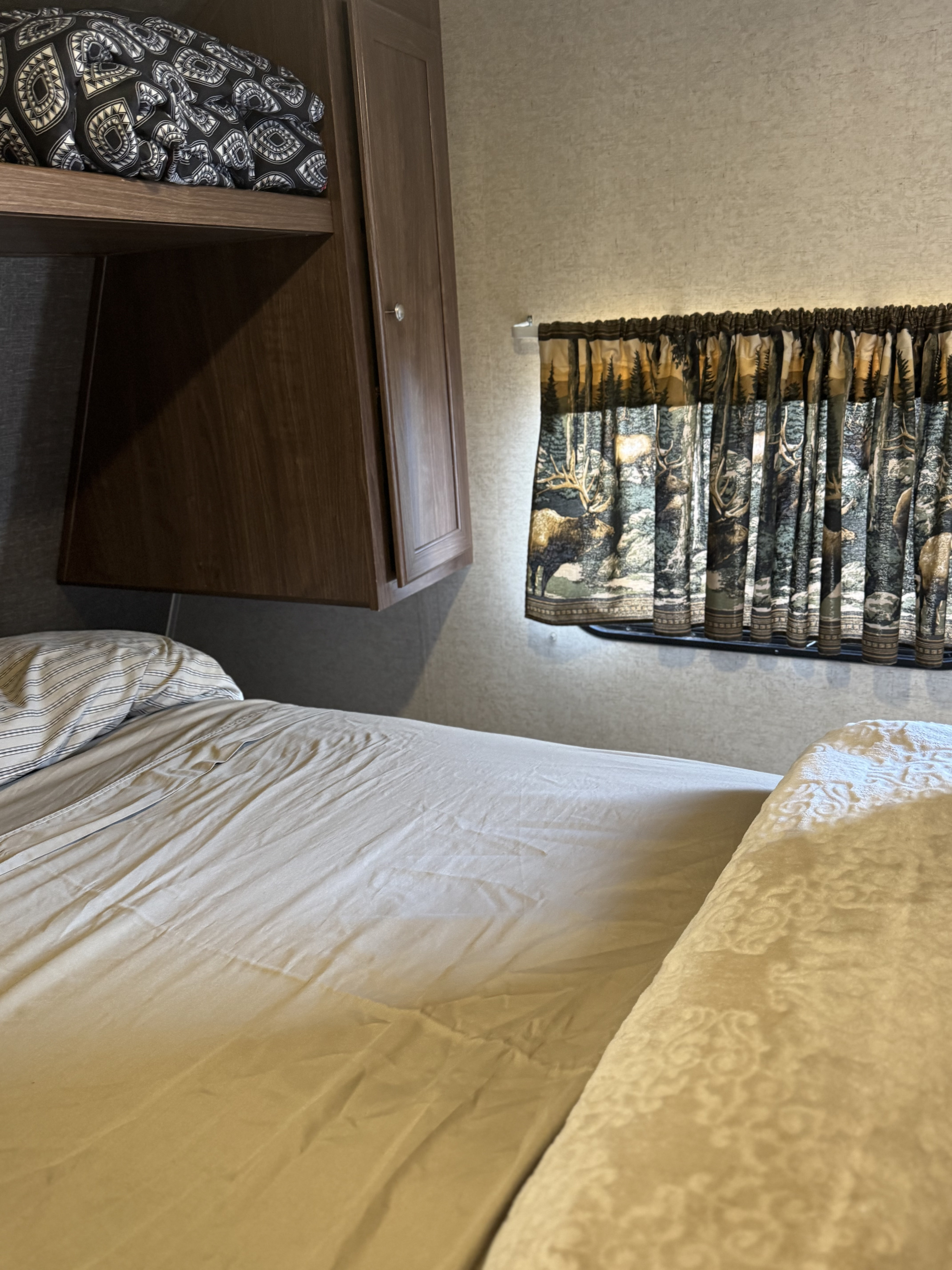 RV rental bedroom with a made bed, wood overhead cabinets, and a window displaying nature-themed curtains.. Heartland RVs Prowler Lynx 2016