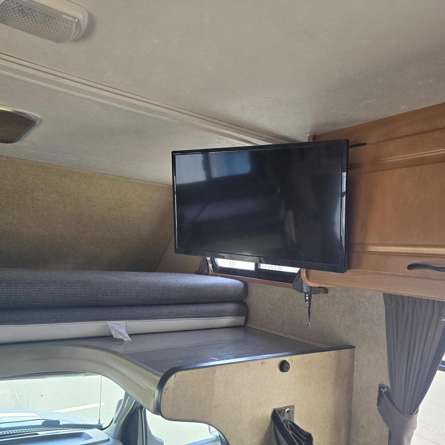 Interior of an RV rental featuring an over-cab sleeping area, a mounted TV, and wood cabinetry.. Thor Majestic 2016