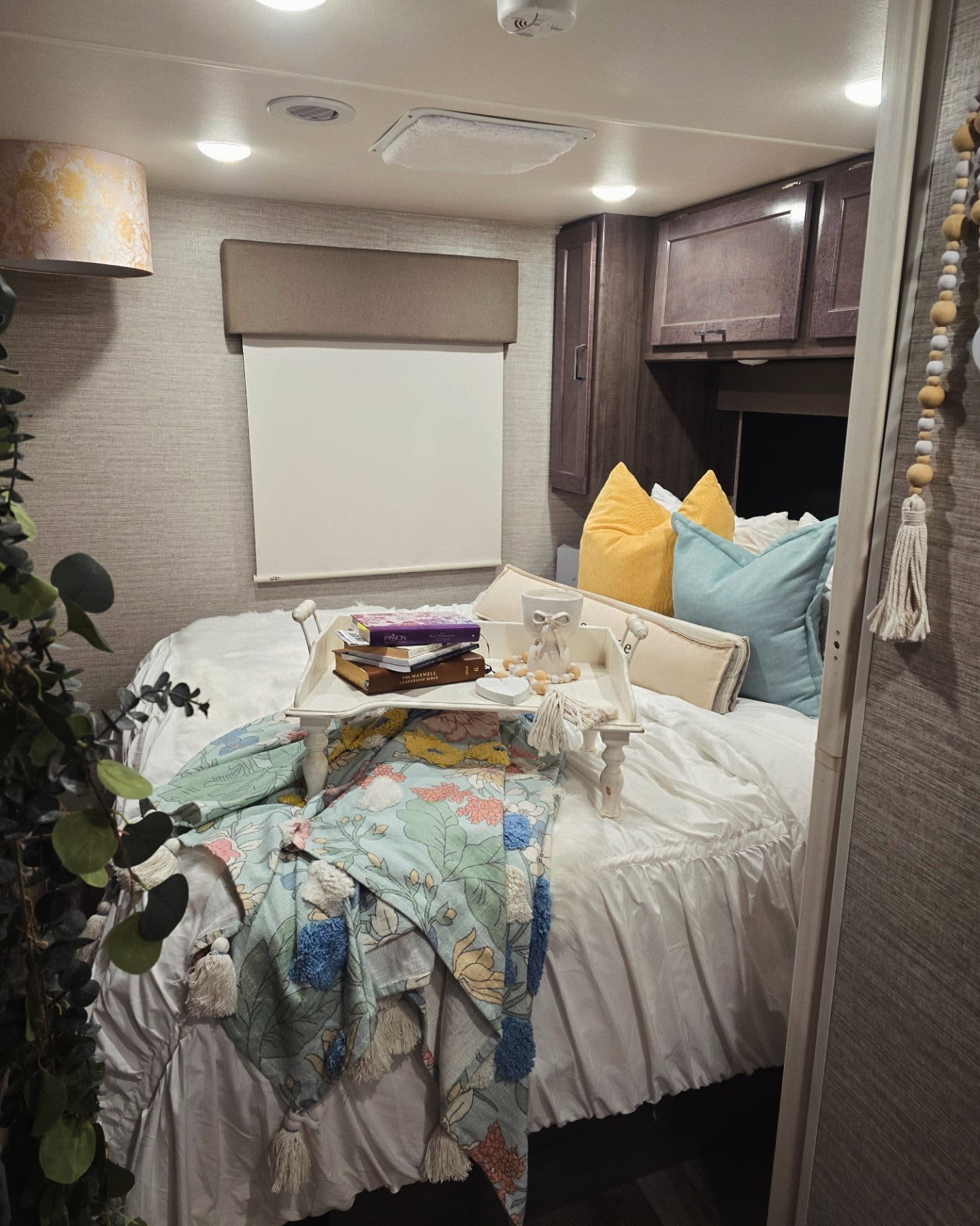 Cozy RV rental bedroom featuring a neatly made white bed with colorful pillows, a floral blanket, and a bed tray with books.. Winnebago Spirit 2024