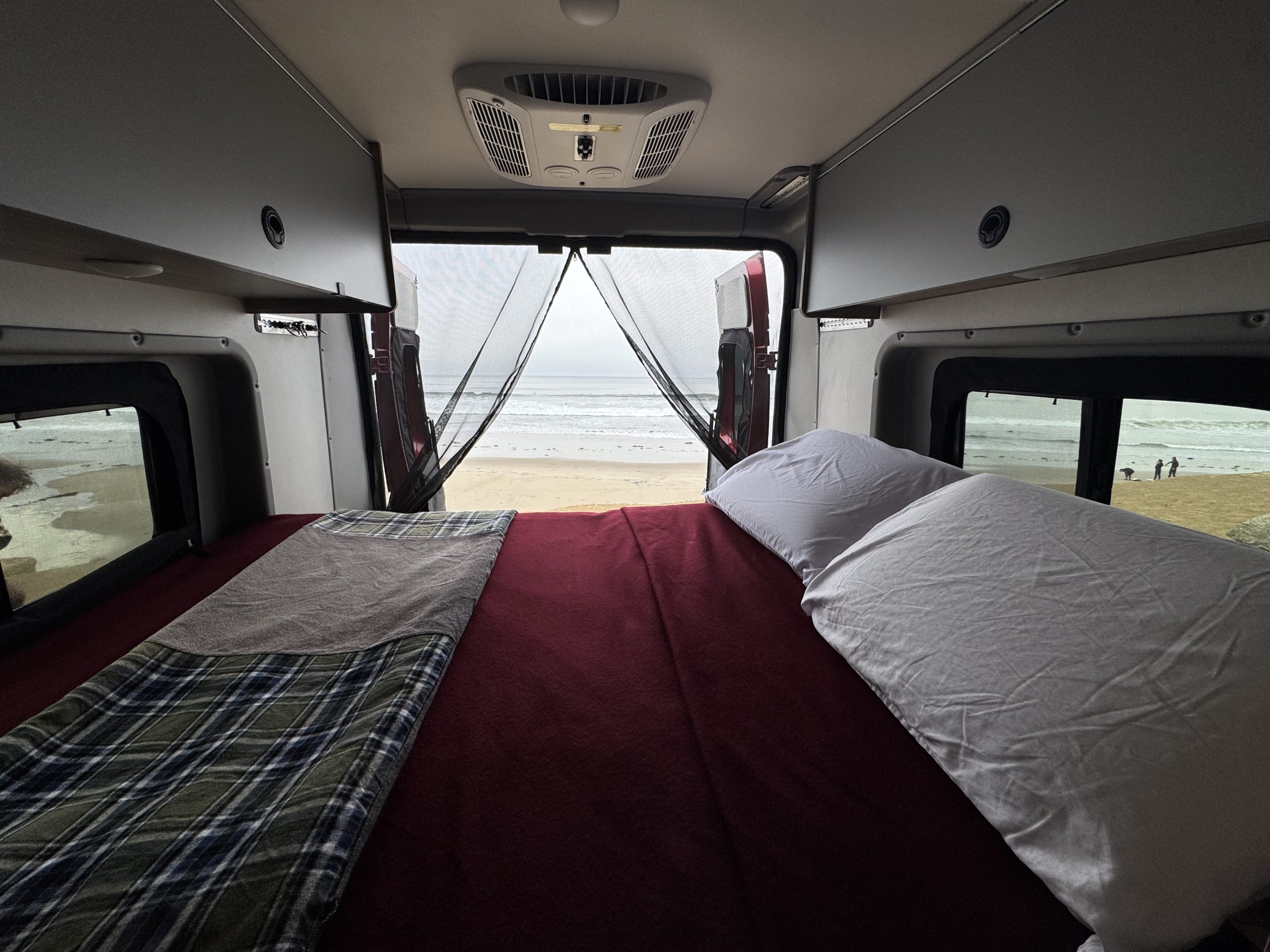RV rental camper van interior. Features a cozy bed with blankets/pillows, and a clear beach/ocean view through open rear doors.. Winnebago Winnebago 2023