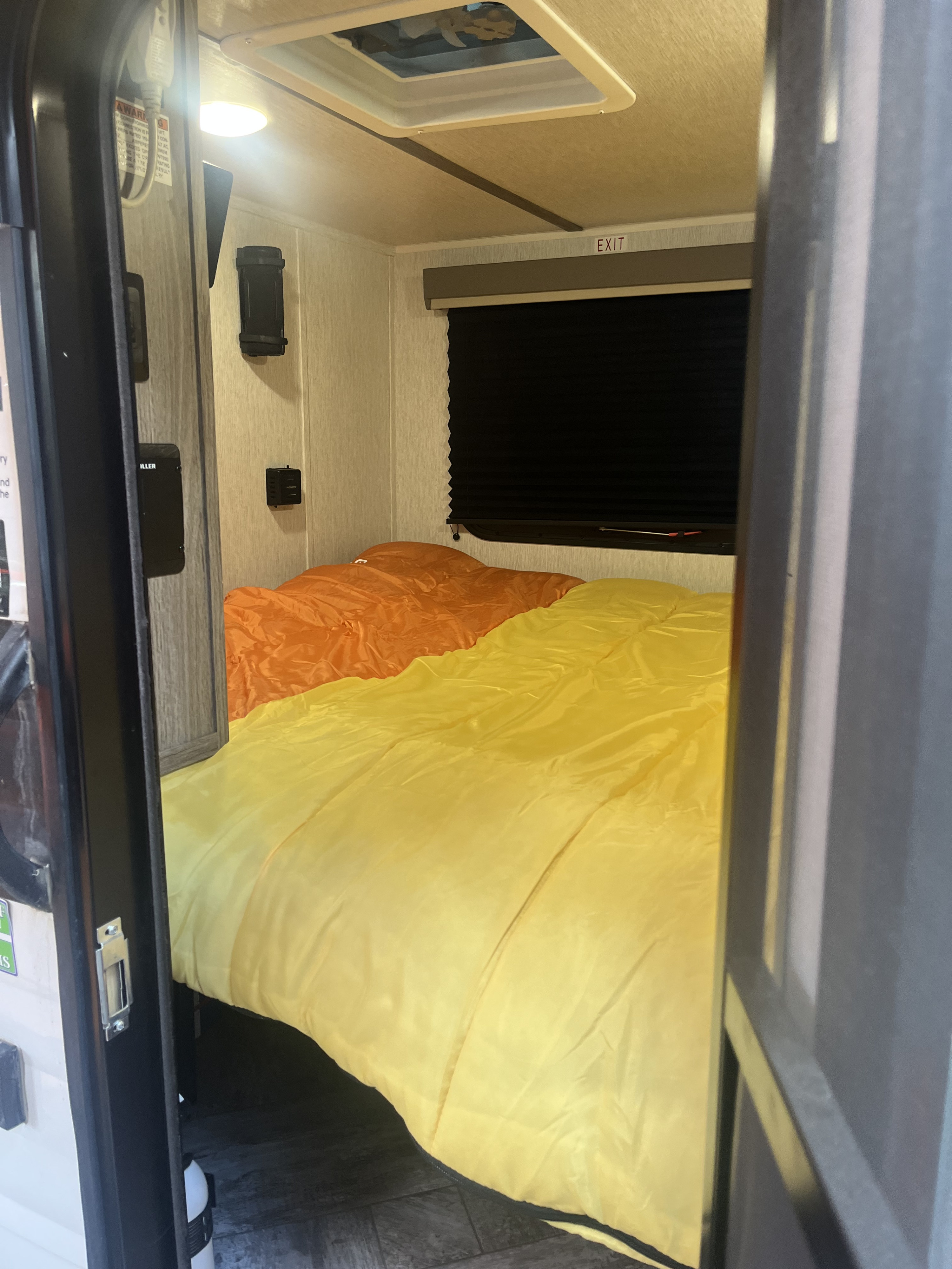 Interior of an RV rental showing a bright orange and yellow bed, window with black blinds, and an overhead exit sign.. Sunset Park RV SunRay 2025