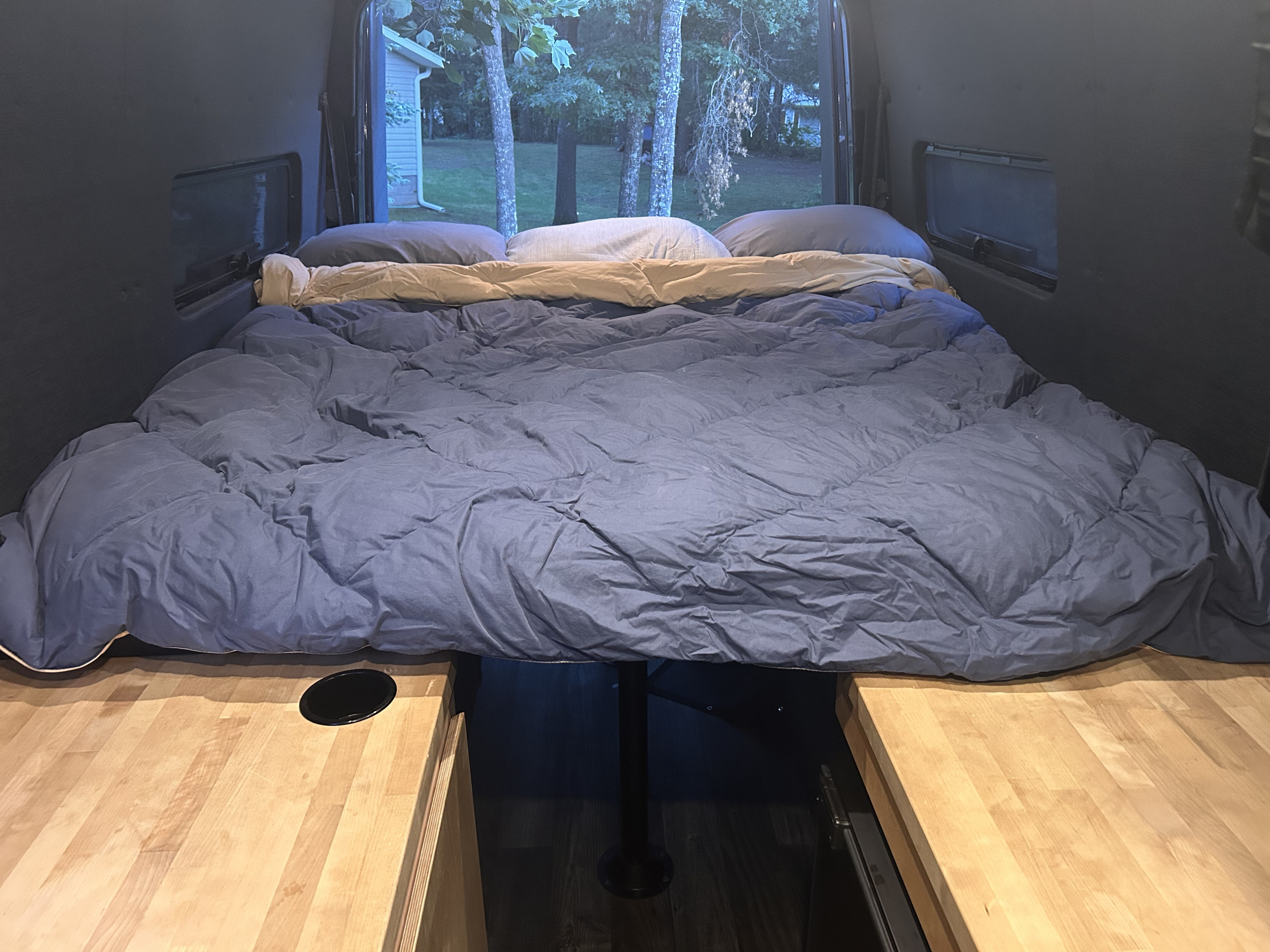 Inside a cozy RV rental: a large bed with blue comforter and pillows, offering a peaceful nature view. Wood counters below.. Mercedes Sprinter 2013