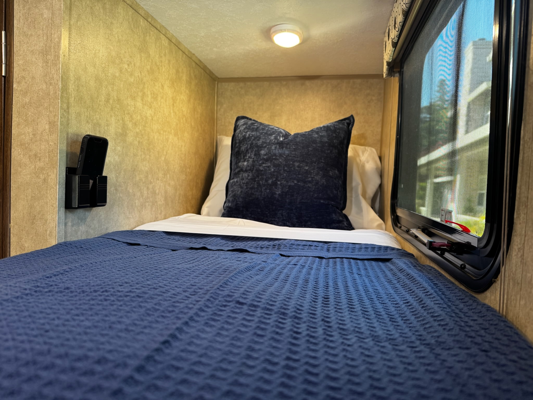 Cozy RV rental bunk with blue bedding, ceiling light, phone holder, and window view.. Clipper Clipper Trailer 2020