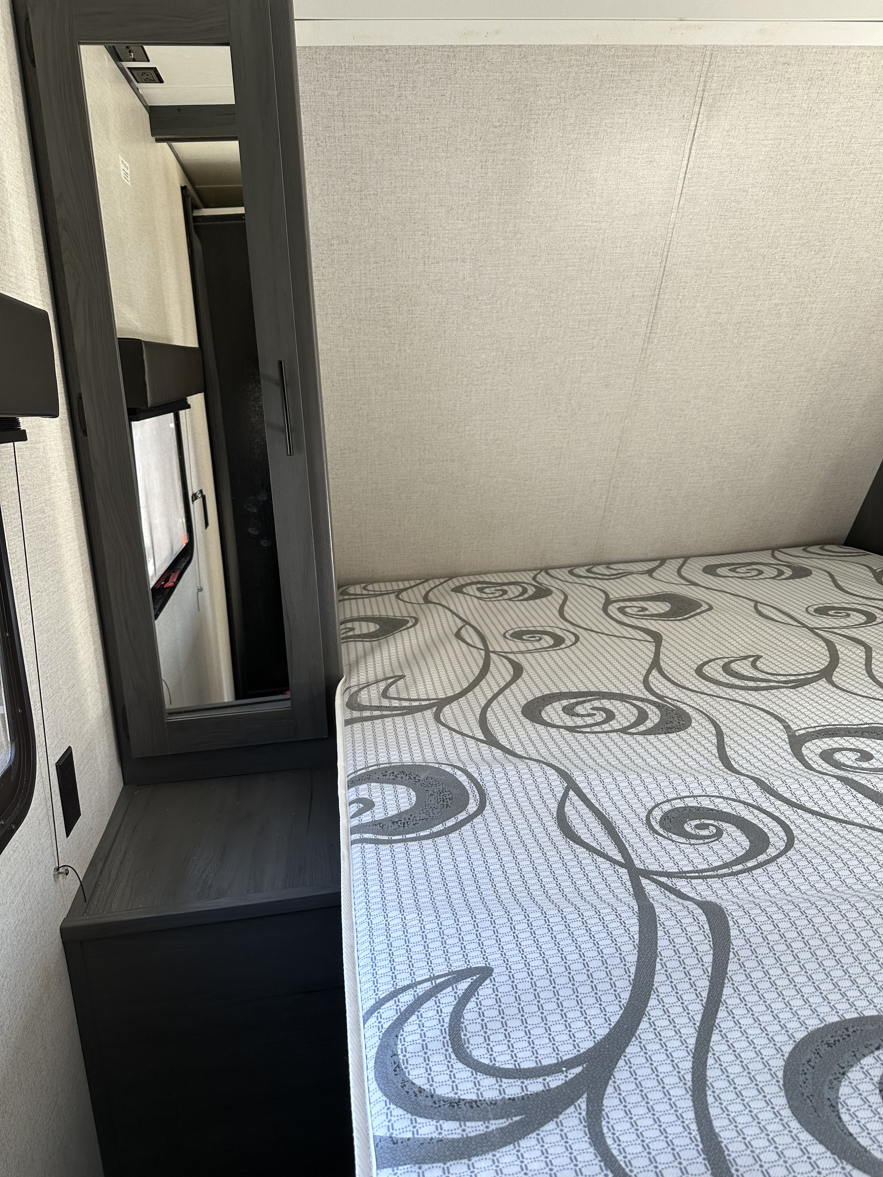 RV rental bedroom with a patterned mattress, dark wood cabinet, and mirror reflecting wall outlets and window.. Dutchmen Aspen Trail LE 29BH 2021