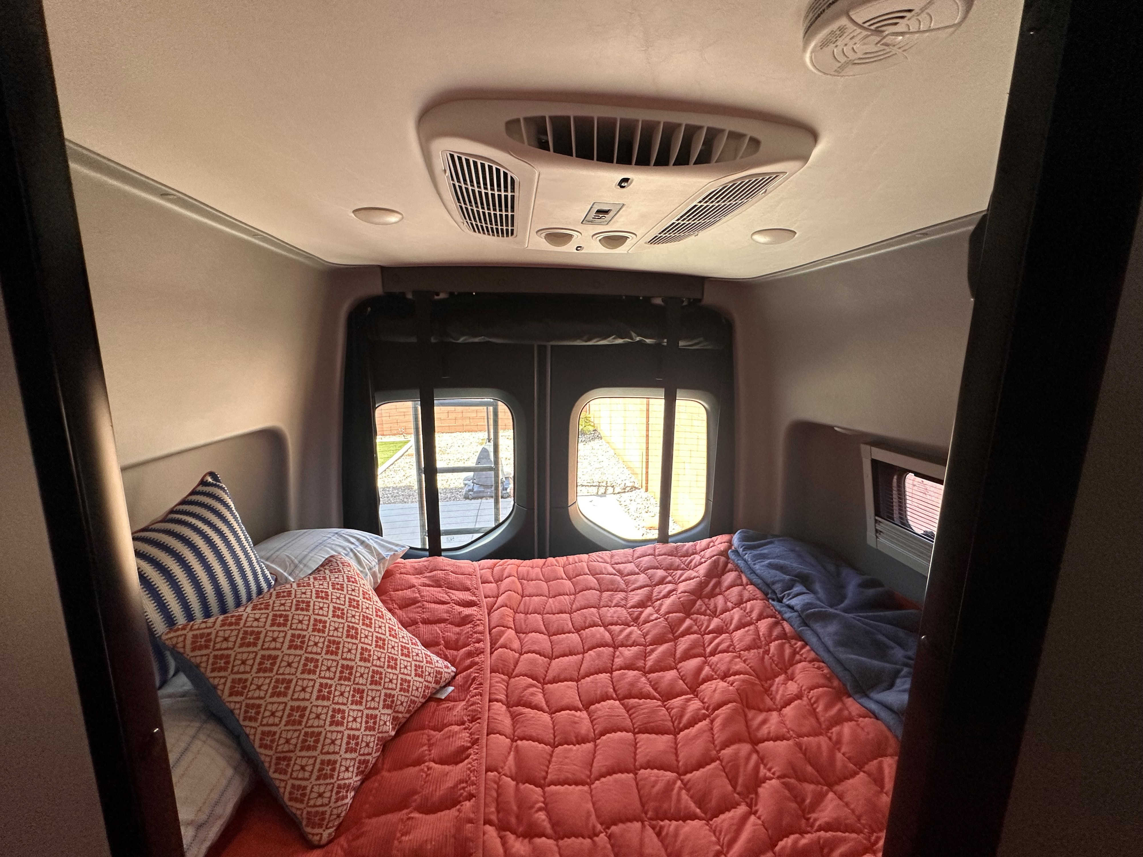 Comfortable RV rental bed setup with a red comforter and pillows. Rear windows show outdoor view; AC unit on ceiling.. Winnebago Revel 2025
