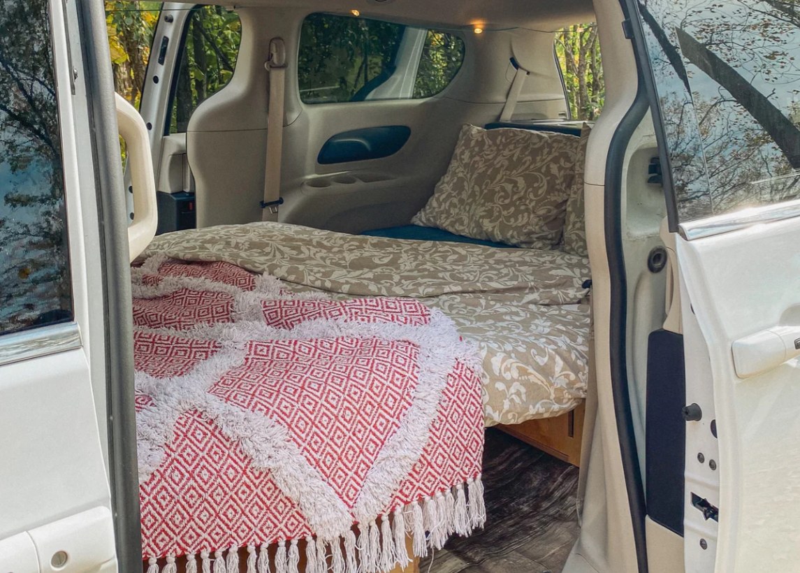 RV rental ready for adventure! Cozy minivan conversion with a comfy bed, beige sheets, and a red fringed blanket.. Dodge B Van 2014