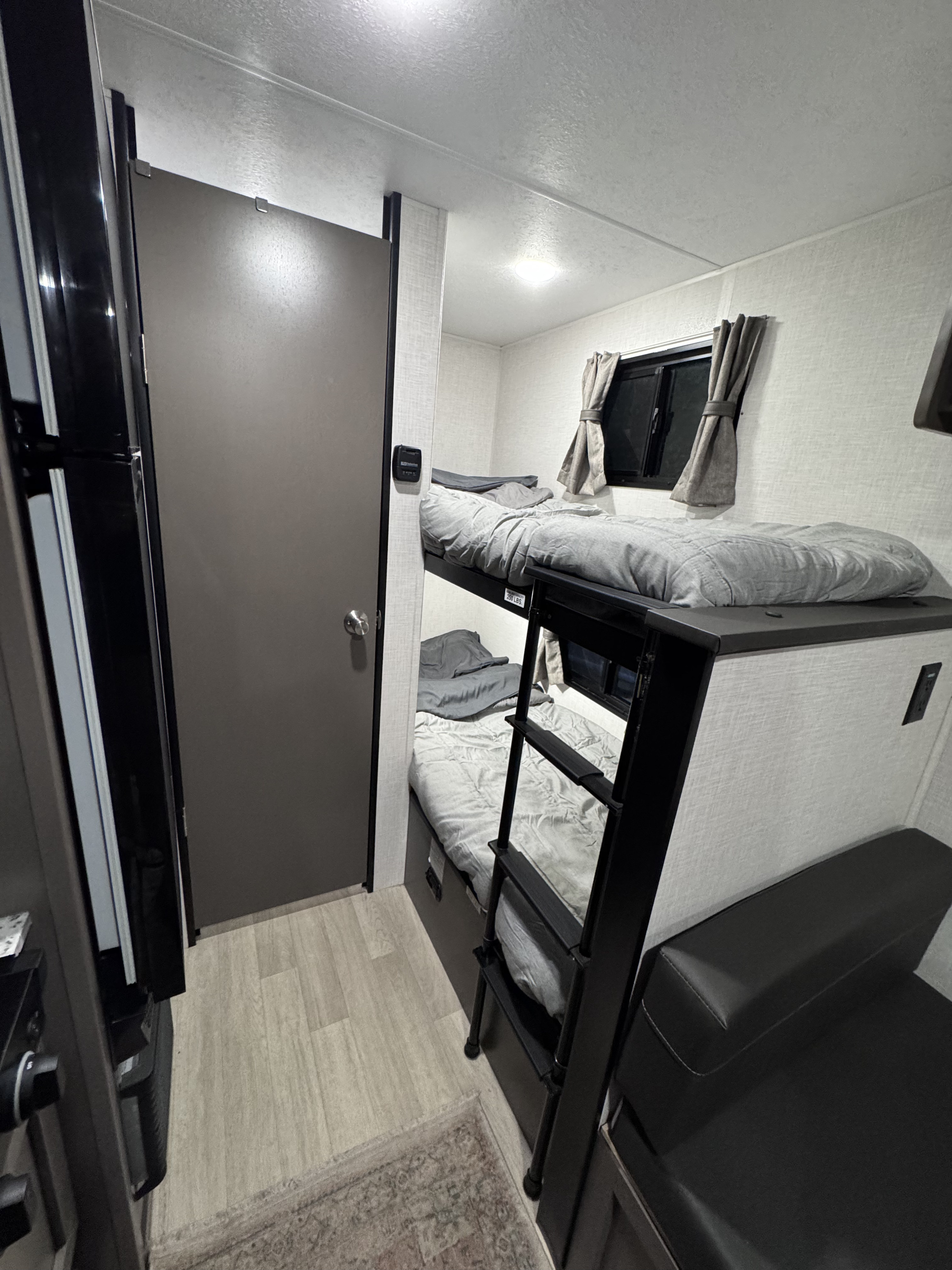 This RV rental features a cozy sleeping area with modern bunk beds, gray bedding, and a side window, ideal for compact living.. KZ Sportsman 2024