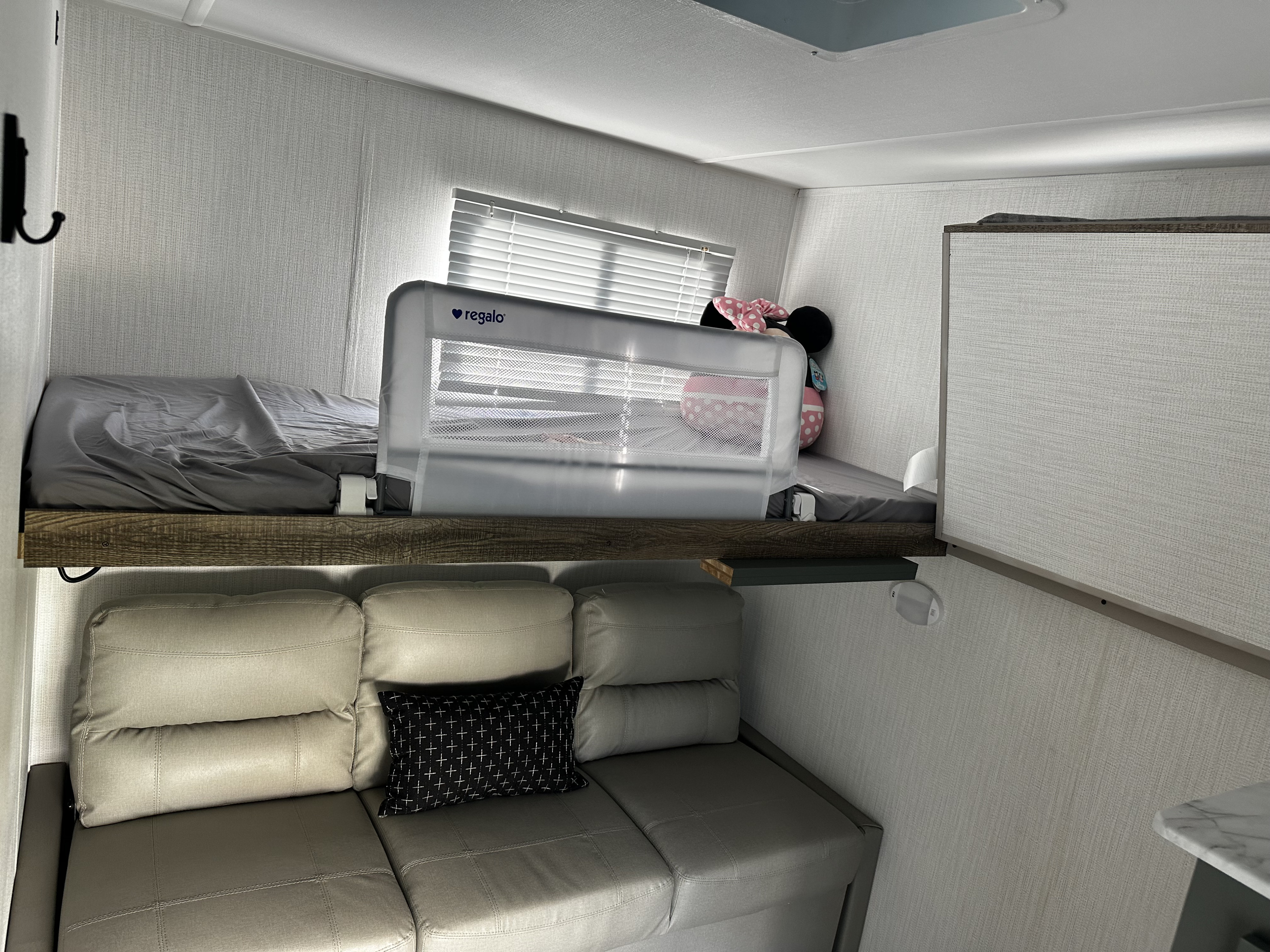 RV rental bunk area: upper bed with a Regalo child safety rail & plush Minnie Mouse, above a tan sofa & window.. Heartland RVs EDDIE BAUER 322BH-C 2024