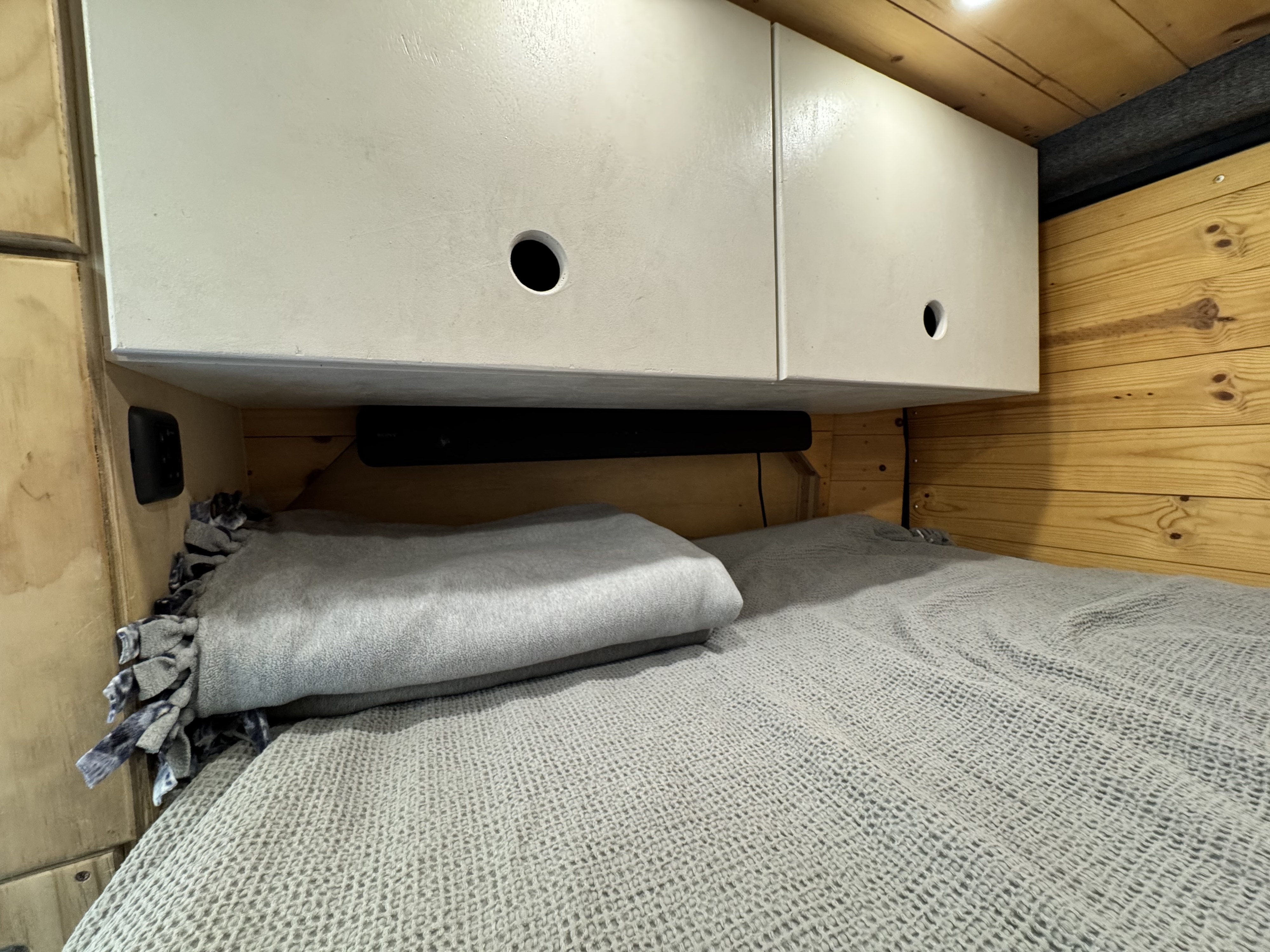 Cozy RV rental bed with overhead cabinets, a soundbar, and warm wood paneling, ready for a comfortable rest.. Ram ProMaster 2500 High Roof 159" WB 2018