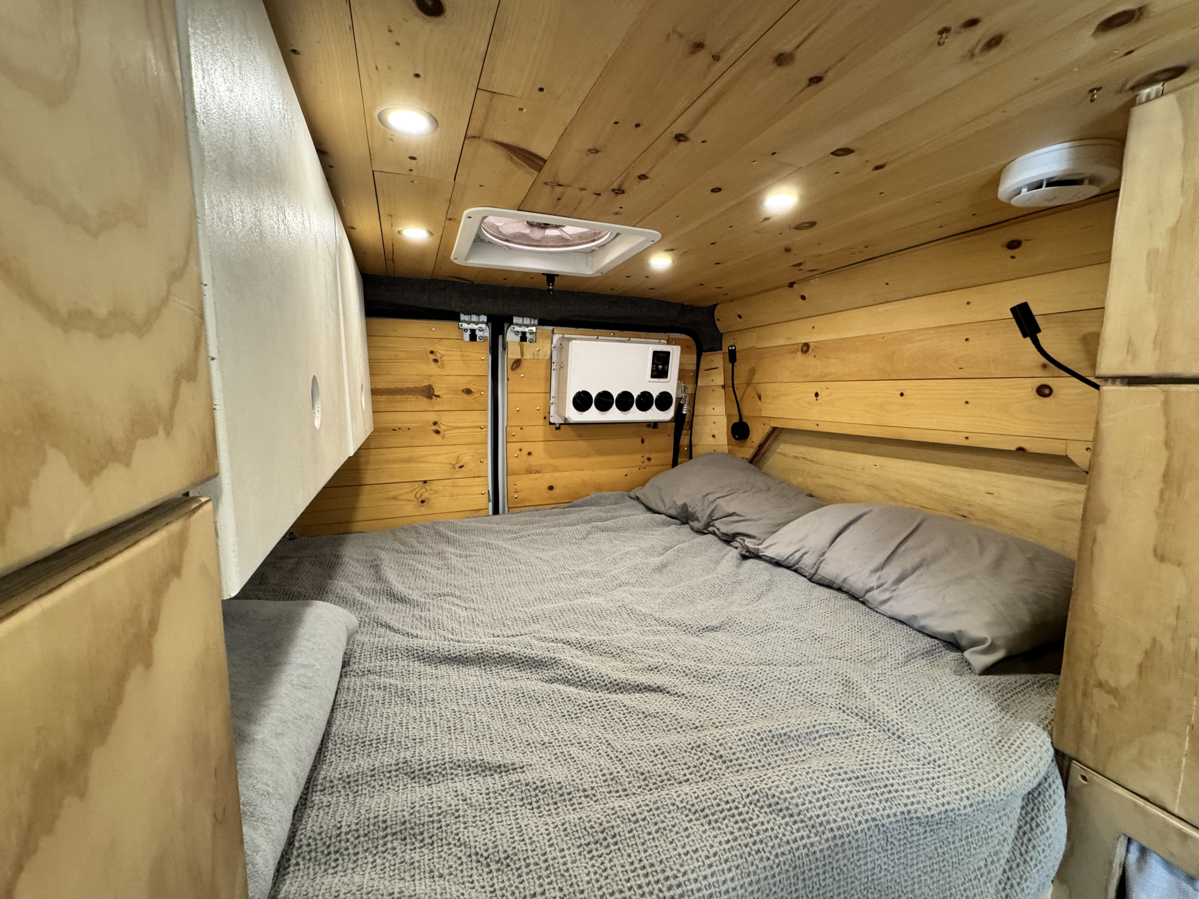 Warm RV rental bedroom with wood paneling, gray bedding, overhead lights, a roof vent, and wall-mounted controls. Left side storage.. Ram ProMaster 2500 High Roof 159" WB 2018