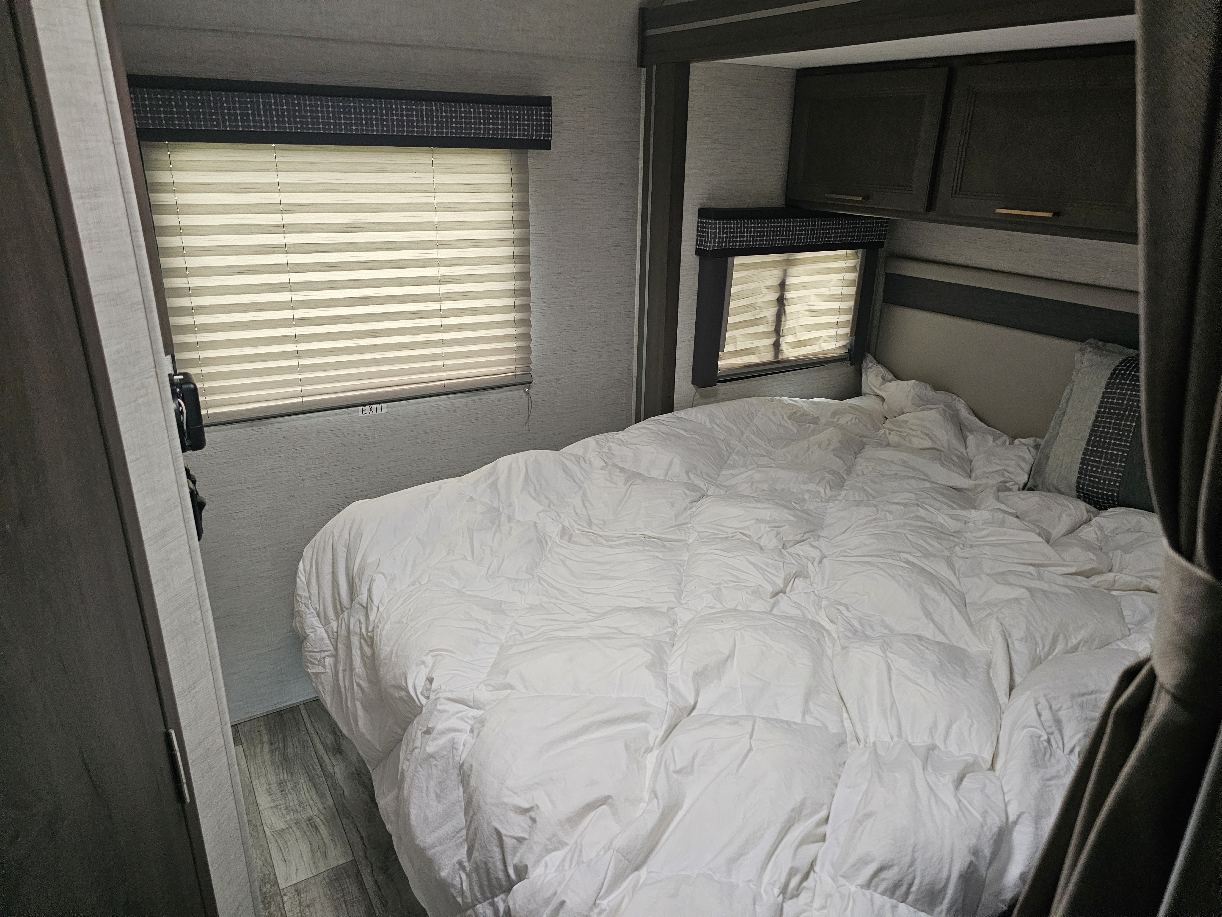 Inside an RV rental bedroom: a large bed with a white comforter, two windows with blinds, dark overhead cabinets, and wood-look flooring.. Thor Chateau 2024