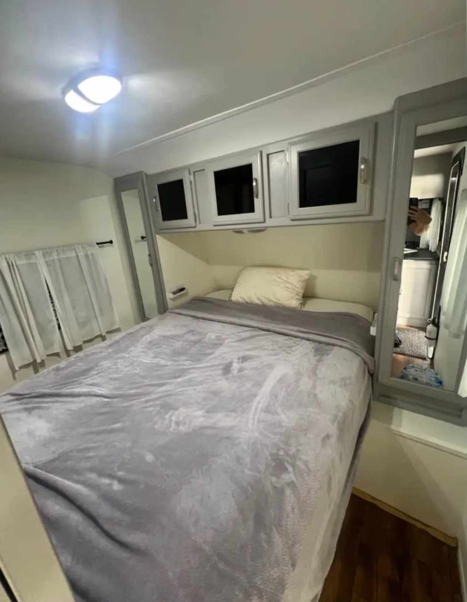 This RV rental features a cozy bedroom: a bed with a grey blanket, overhead cabinets, and mirrors reflecting a hallway.. Keystone RV Passport Grand Touring 2015