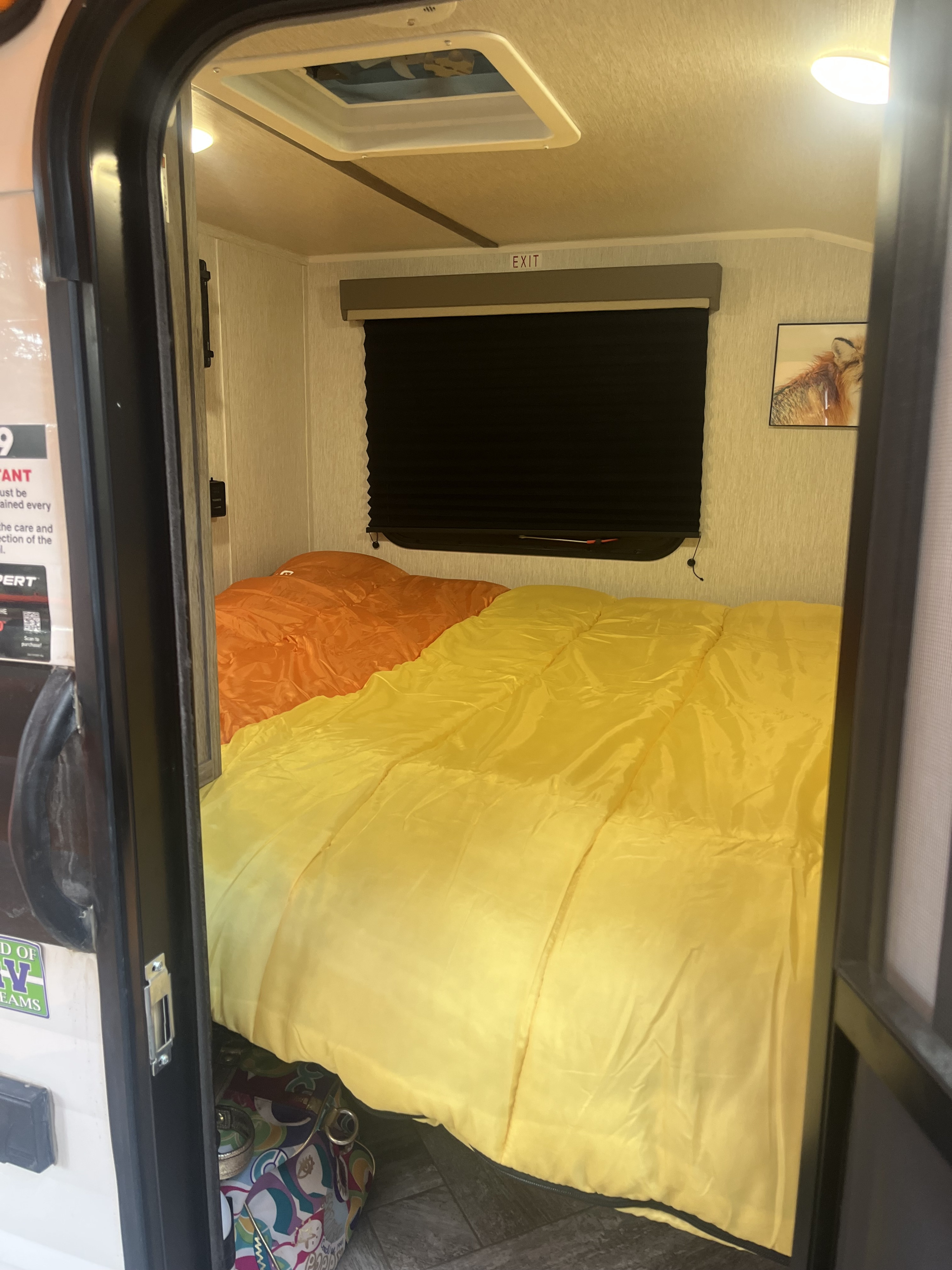 A vibrant RV rental interior, showing a bright yellow & orange bed, black window blind, and overhead vent.. Sunset park rv Sunray 109 2025