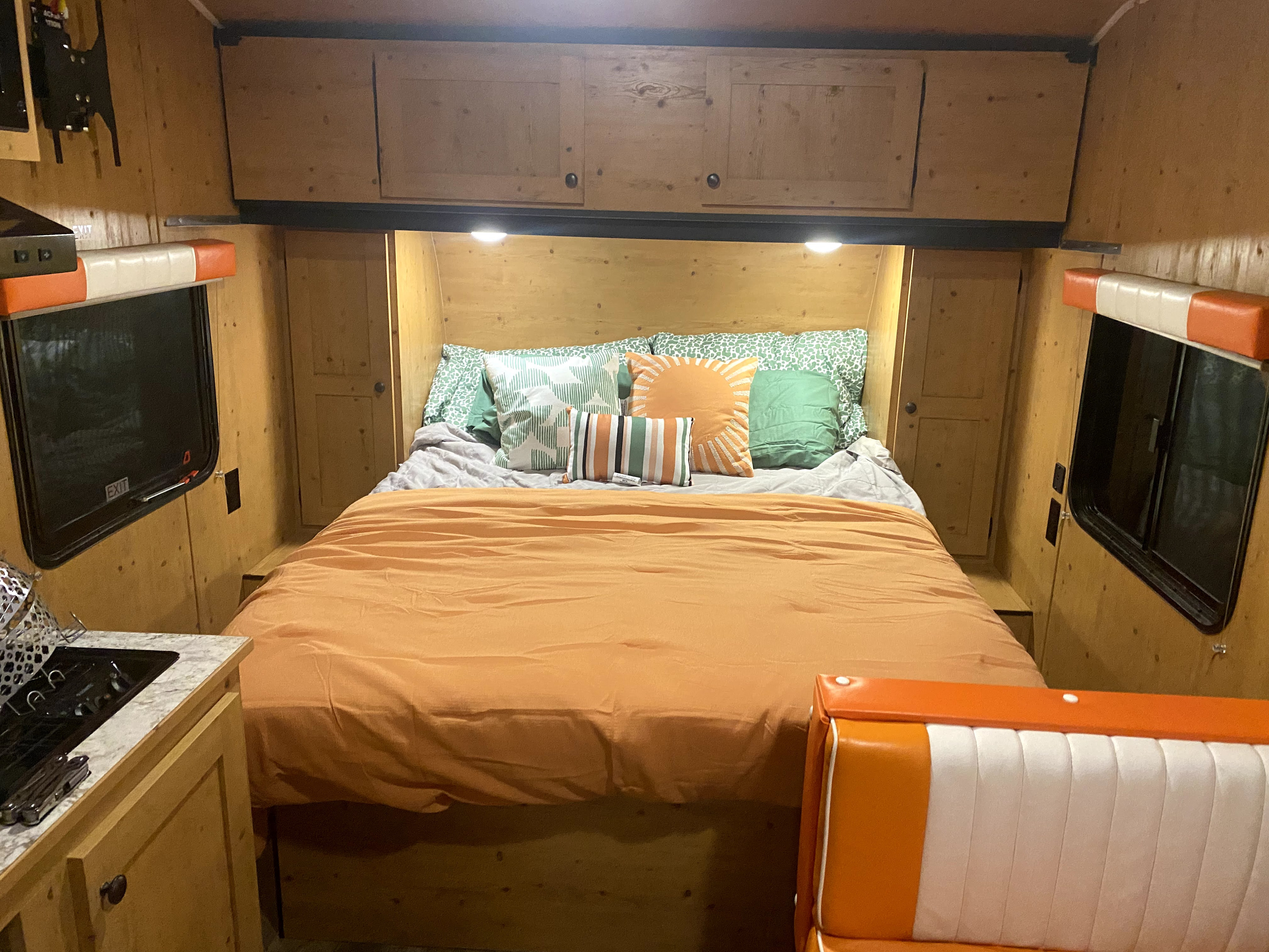 Cozy RV rental interior: wood-paneled bedroom with a bright bed, a compact kitchen, and a retro orange and white dining booth.. Sunset Park RV Sun Lite 2020