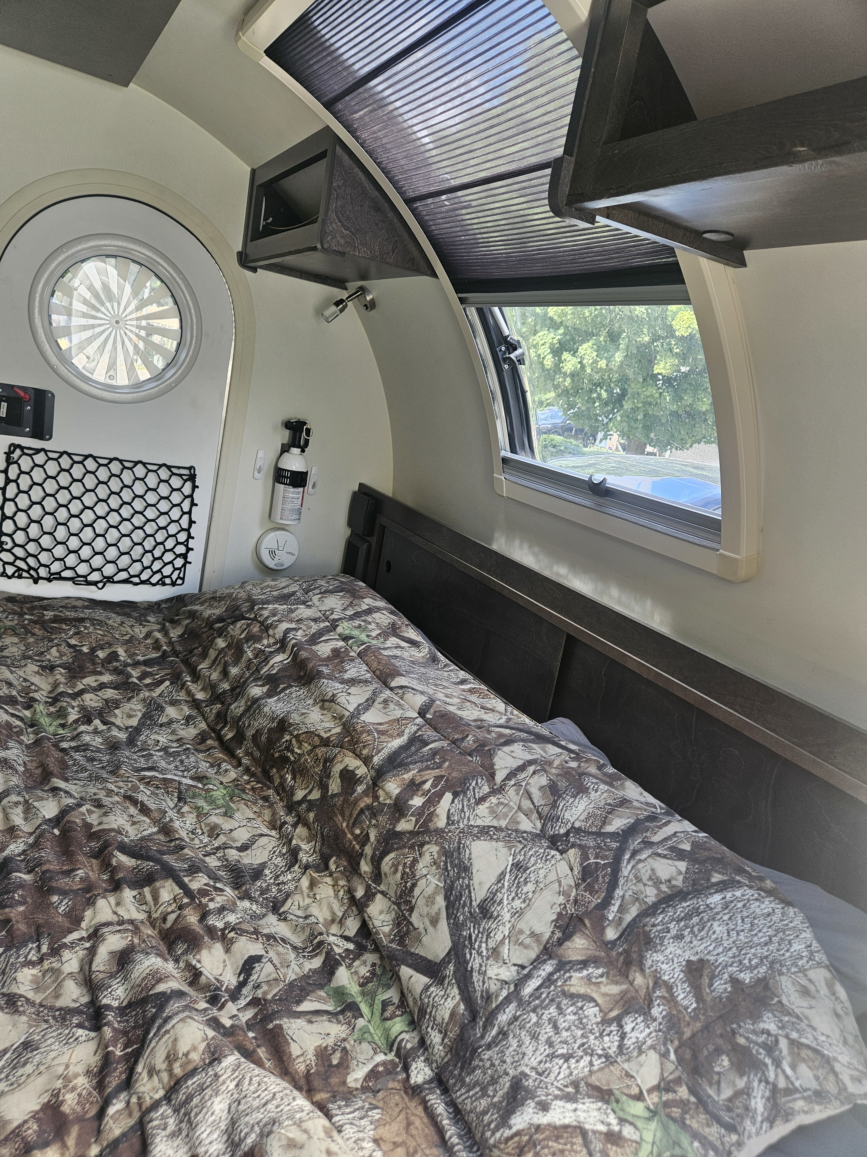 Cozy RV rental interior: camo bed, round entry door, side window with pleated shade, & overhead storage for compact comfort.. nuCamp T@G 2021