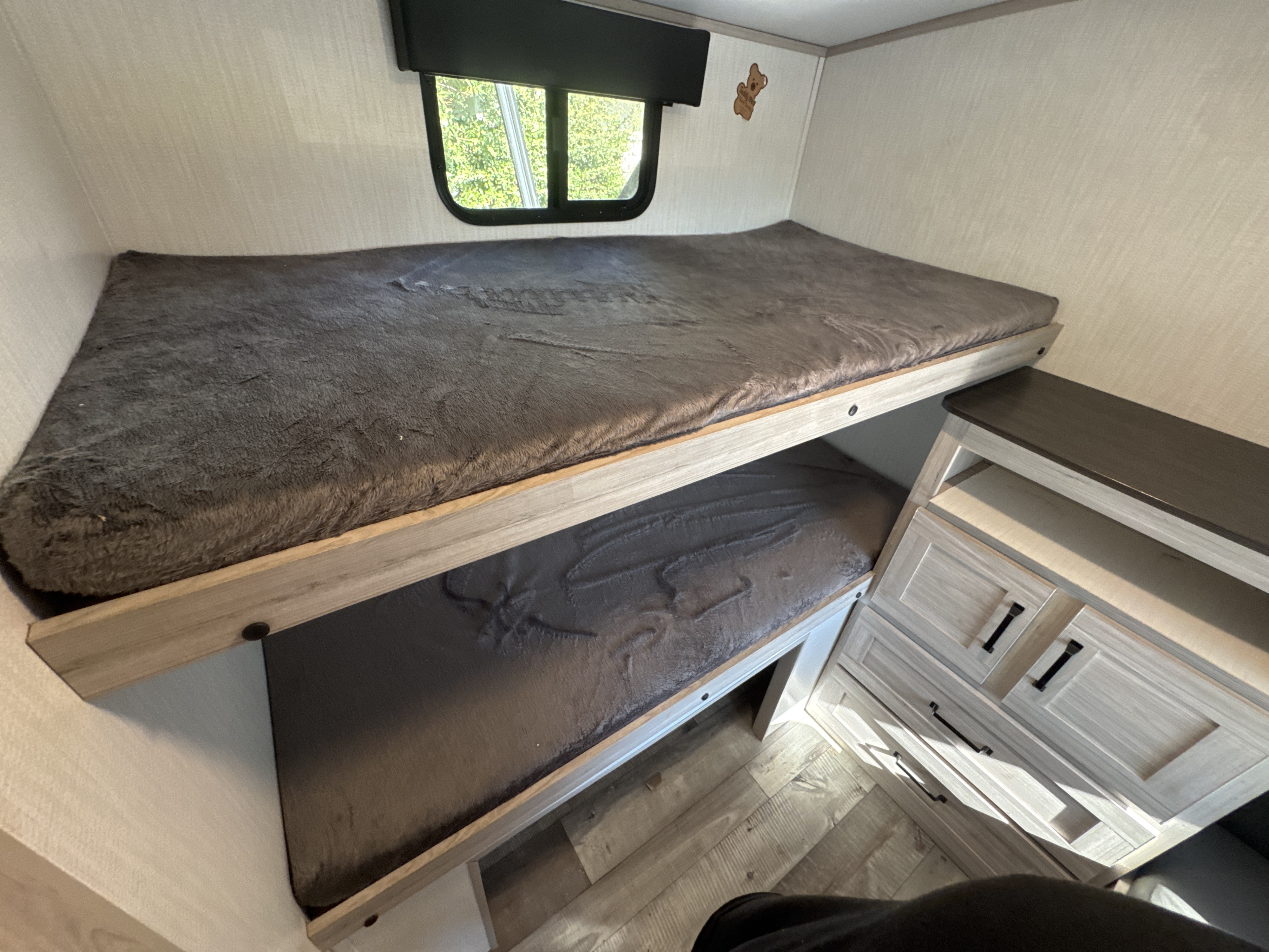 An RV rental interior featuring stacked bunk beds with plush grey bedding, a window, and a storage dresser.. Heartland RVs Sundance Ultra Lite 2024