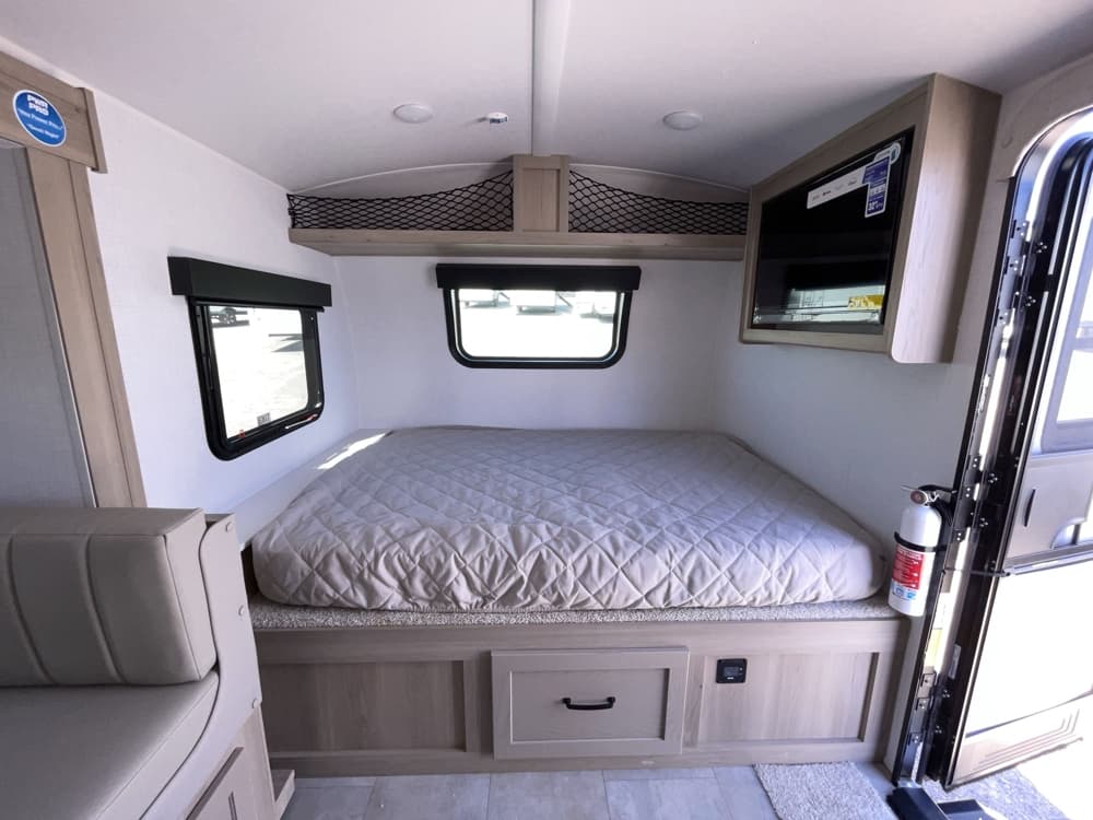 A compact RV rental interior featuring a bed, side windows, overhead storage, and a wall-mounted TV. Ready for your next trip.. Forest River RV Rockwood GEO Pro 2025