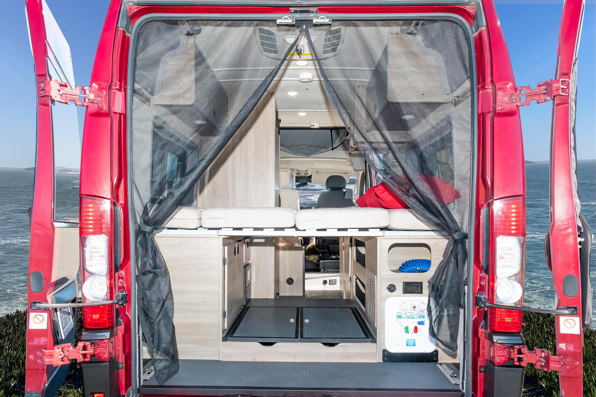 Explore this RV rental's spacious interior, revealed through open rear doors with a protective mesh screen, set against a stunning ocean vista.. Winnebago Solis 2023