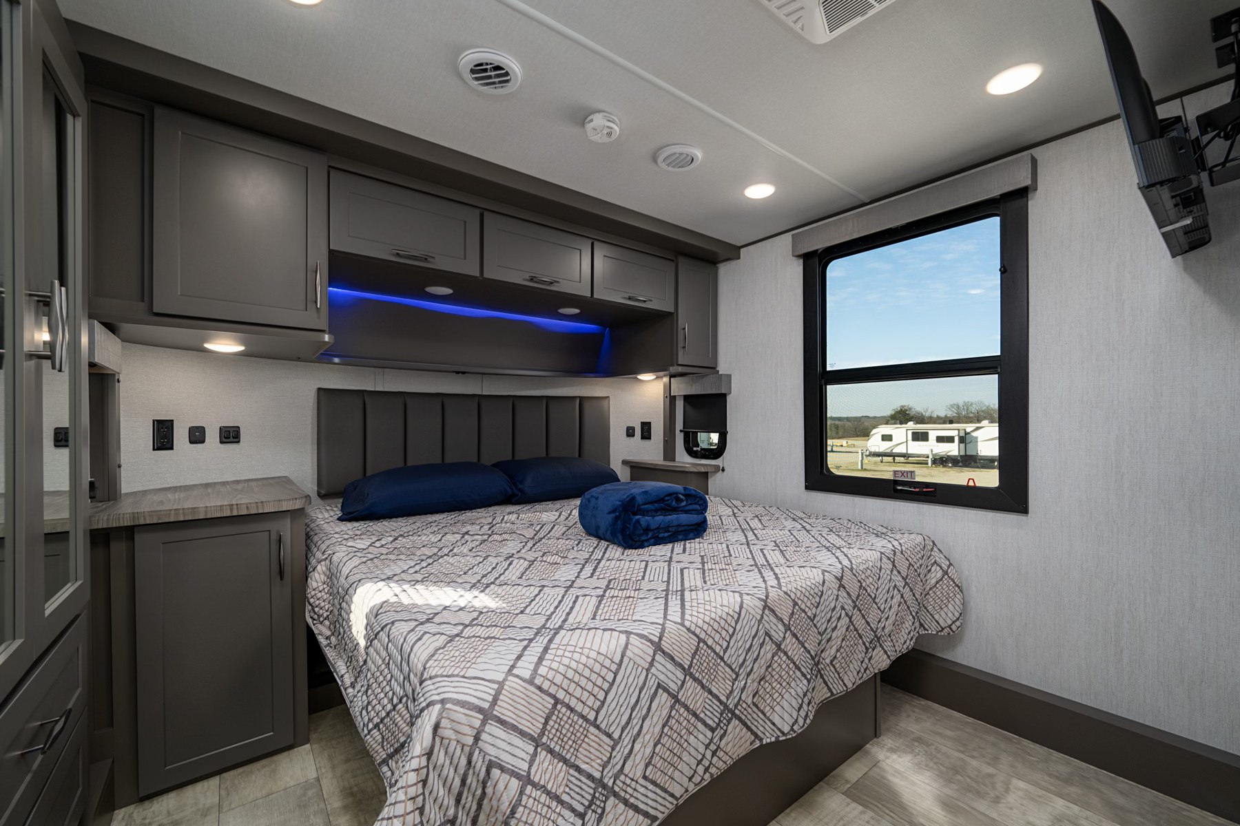 Explore this inviting RV rental bedroom with a comfy bed, sleek gray cabinets, blue accent lights, and wall TV.. Grand Design Momentum 2024