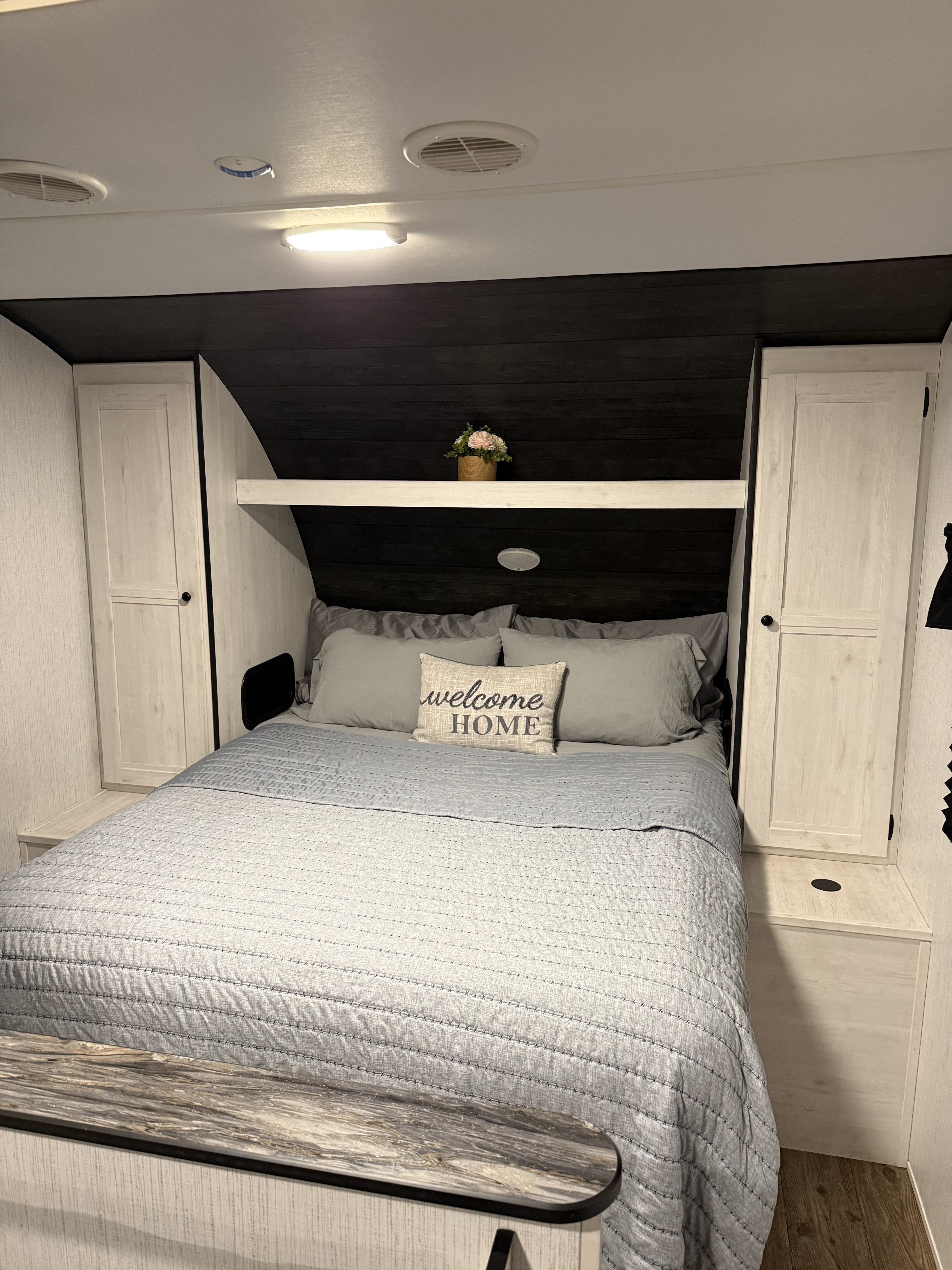 Interior view of an RV rental bedroom: light gray bed with "welcome HOME" pillow, surrounding white cabinets, and dark wood accents.. Heartland RVs Trail Runner 2022