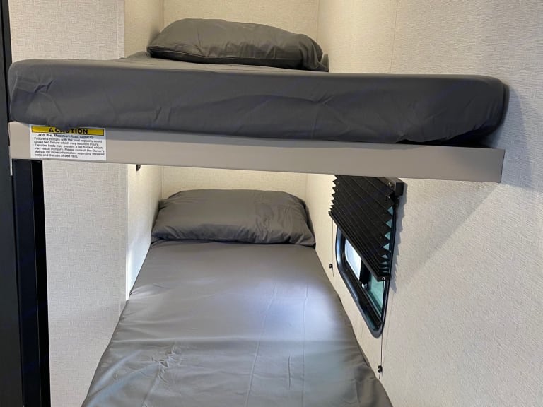 Inside an RV rental, two bunk beds with grey bedding offer compact sleeping. Note the top bunk's weight limit label and window below.. Keystone RV Springdale 2024