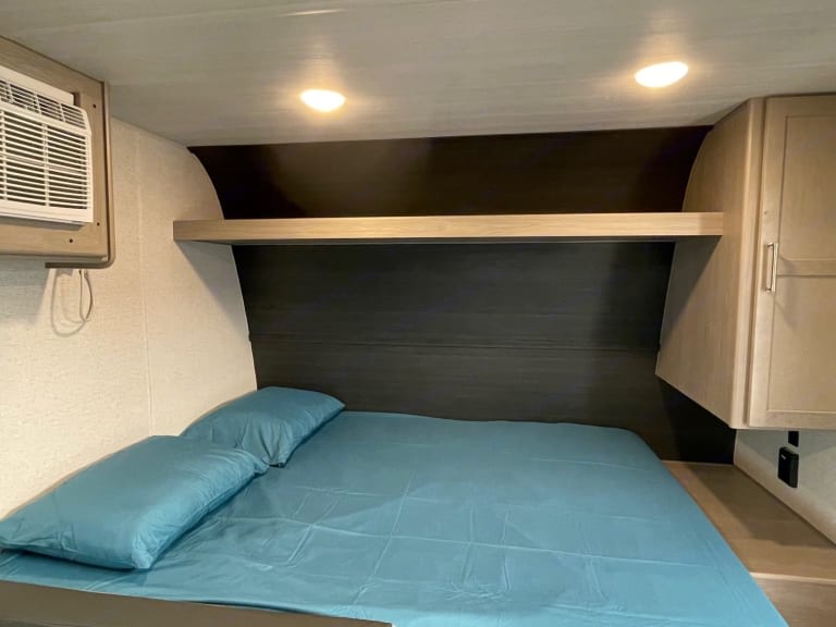 Comfortable RV rental bedroom: blue sheets on bed, overhead shelf, AC unit, and built-in storage for travel.. Keystone RV Springdale 2024