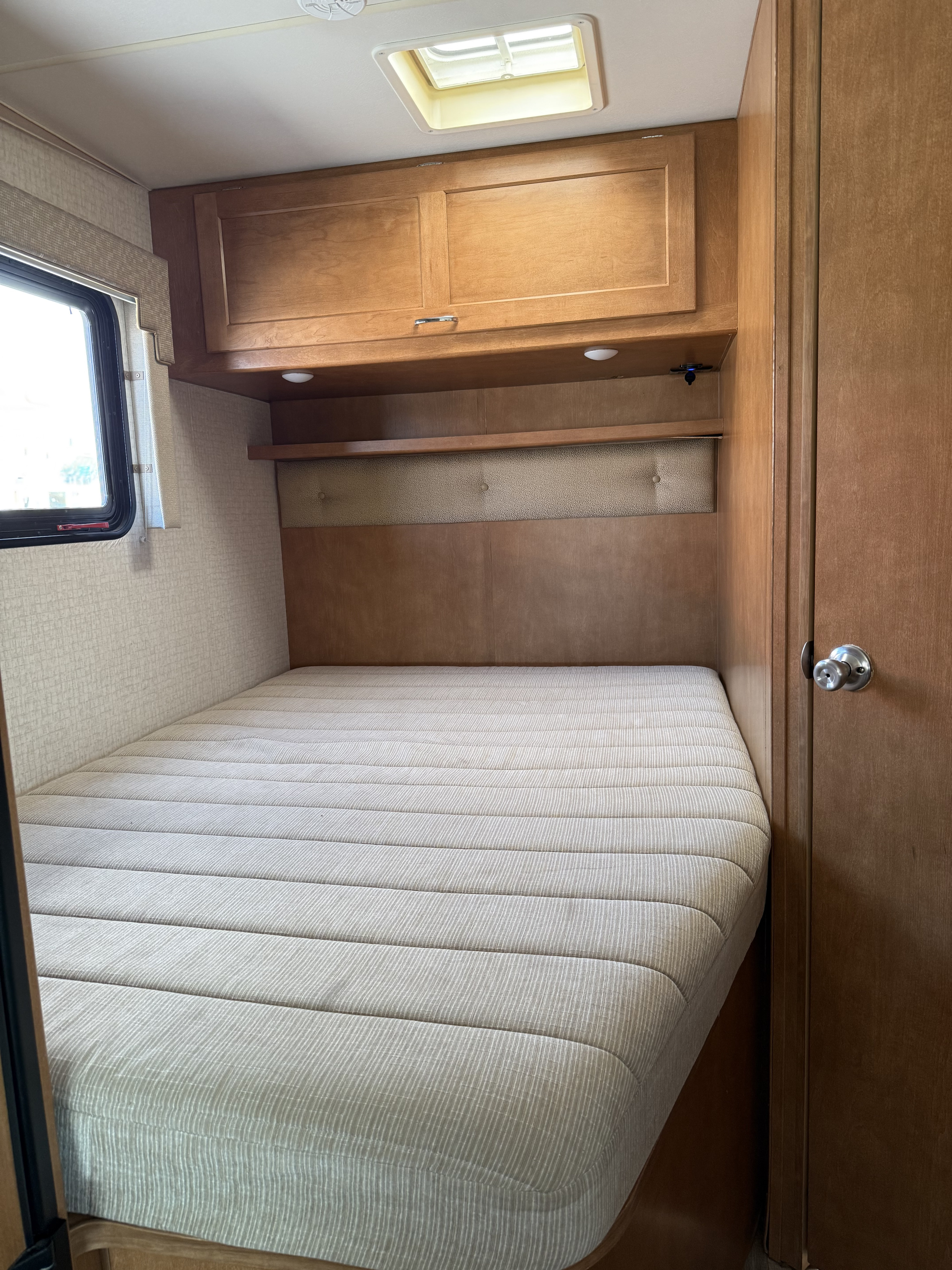 RV rental bedroom with a striped mattress, overhead wood cabinets, and window, providing a cozy sleeping space.. Winnebago Minnie Winnie 2016