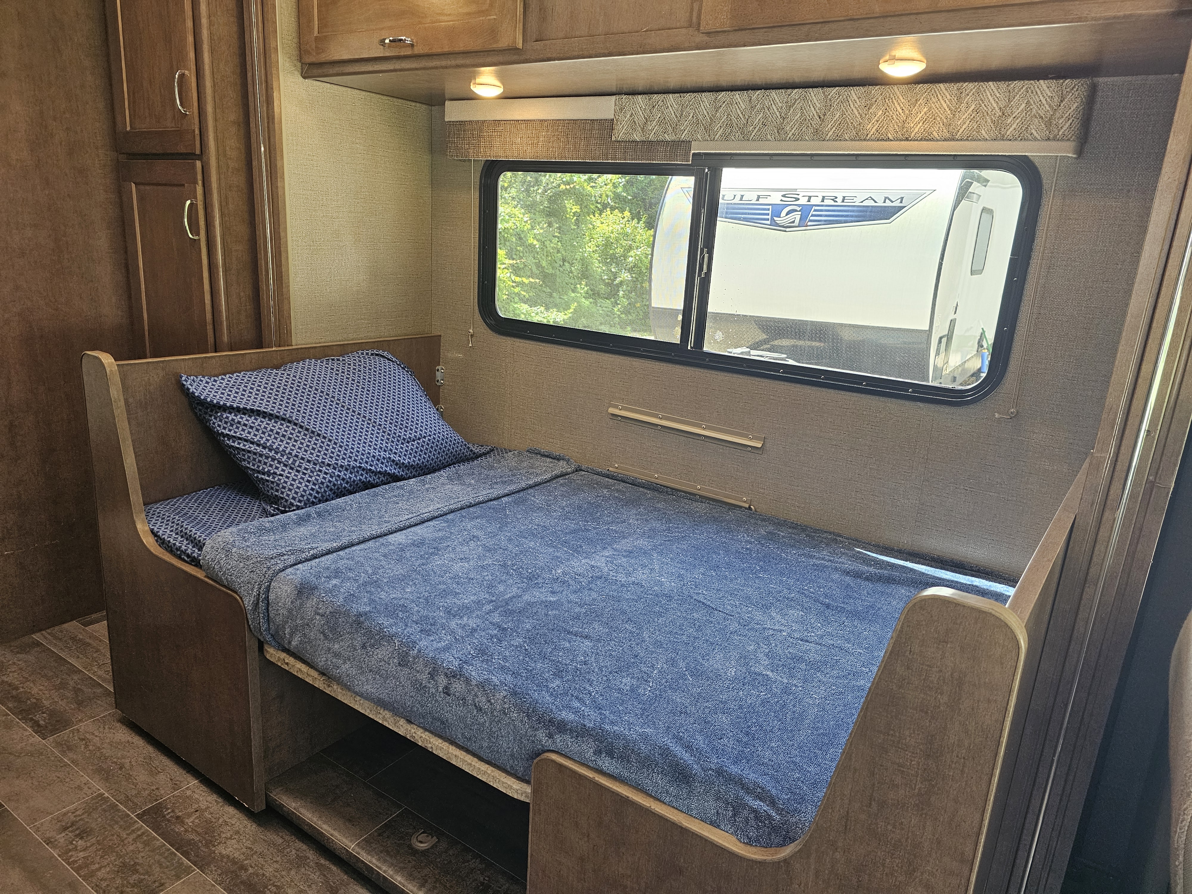 Cozy RV rental bedroom featuring a bed with blue bedding, a window overlooking trees, and light wood cabinetry.. Winnebago Outlook 2019