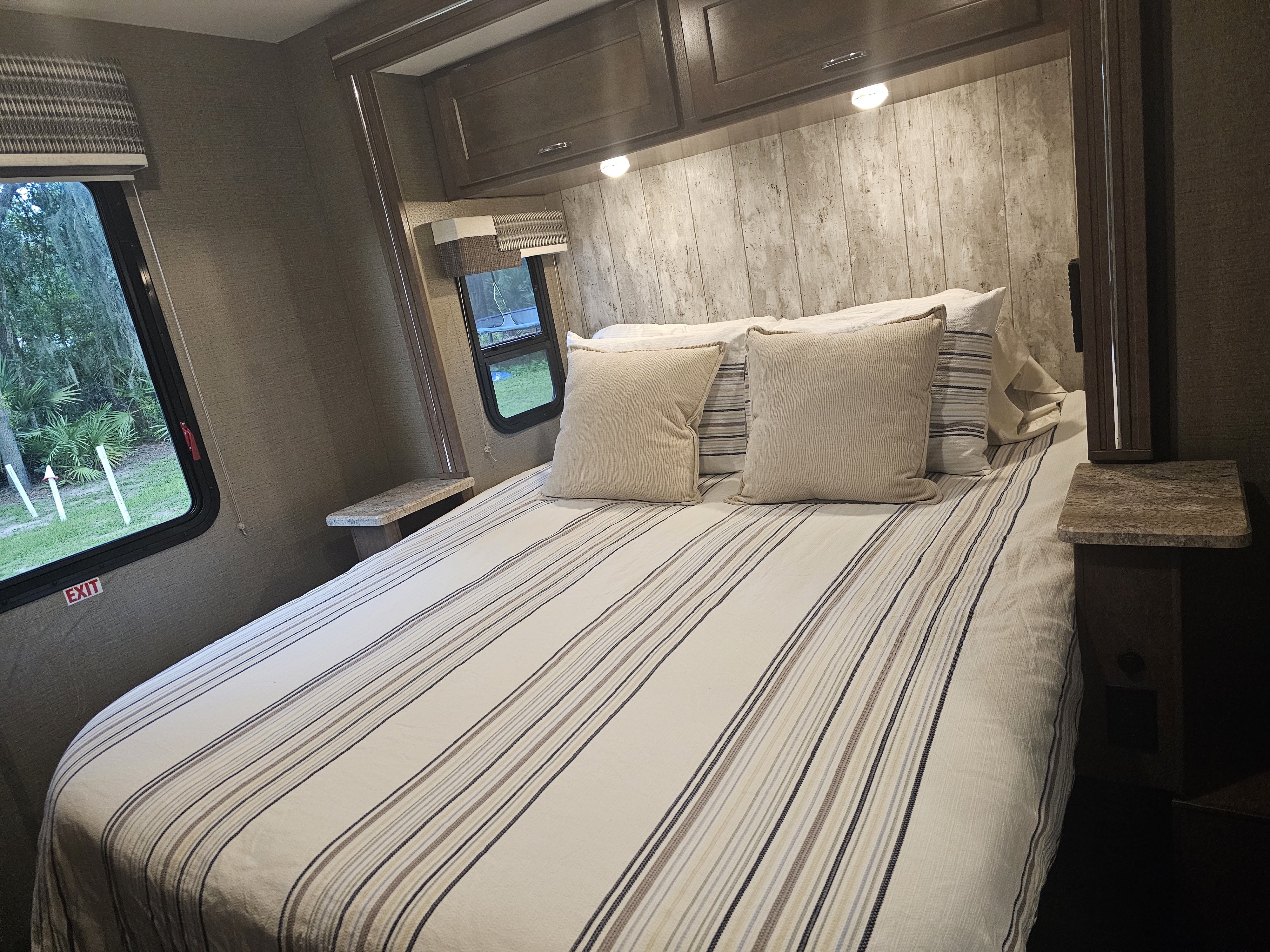 Comfortable RV rental bedroom featuring striped bedding, overhead cabinets, and a window view of nature for your next trip.. Winnebago Outlook 2019