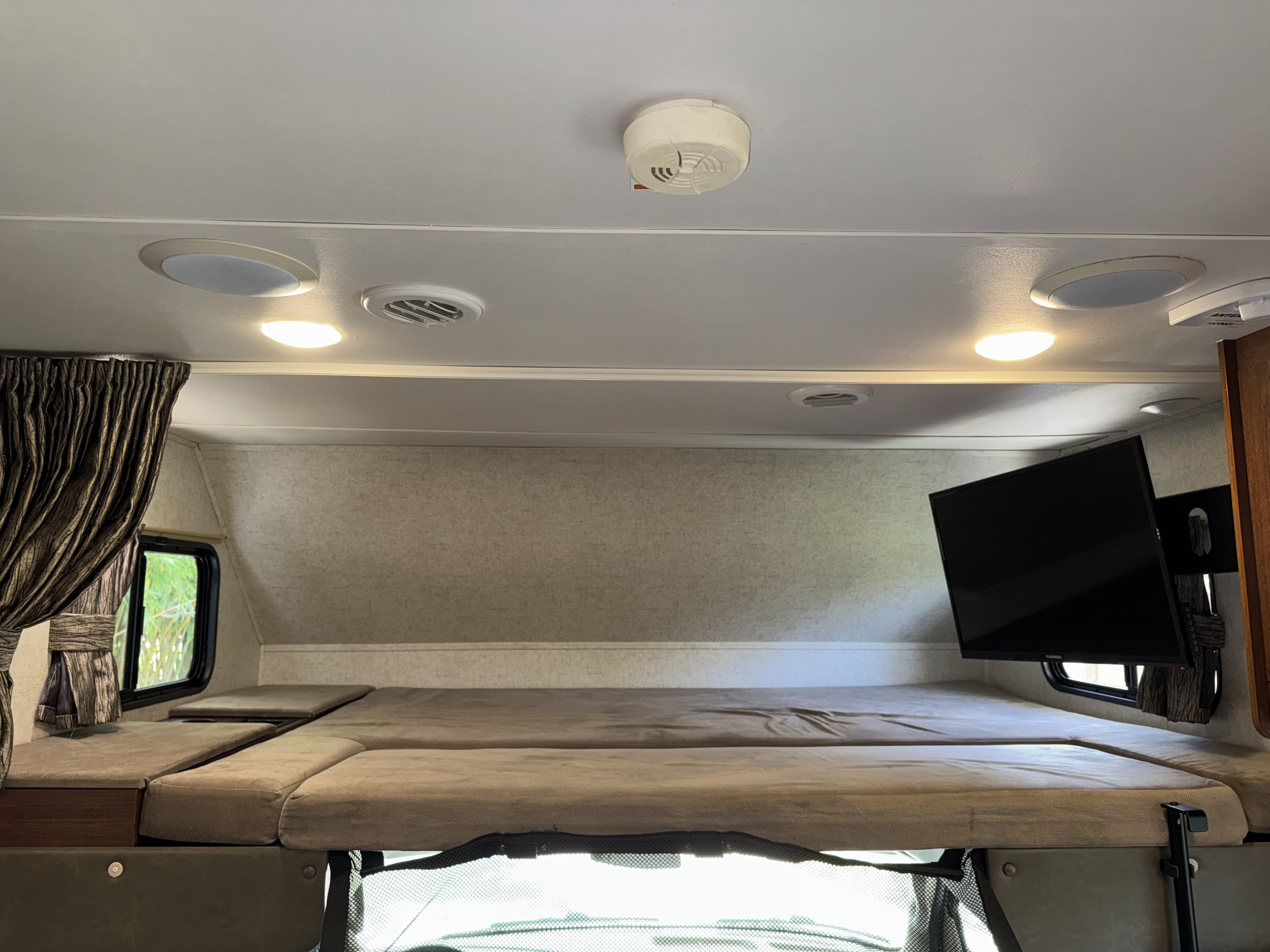 Interior of an RV rental: a cozy cab-over bunk bed, mounted TV, ceiling lights, speakers, and window view.. Forest River Sunseeker 2017