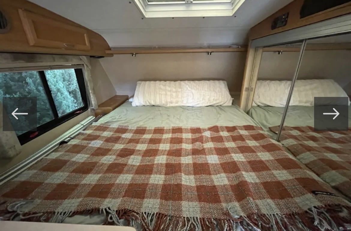 Cozy RV rental bedroom with a comfy bed, plaid blanket, window view of trees, and mirrored storage, ideal for travel.. Lance 951 2007