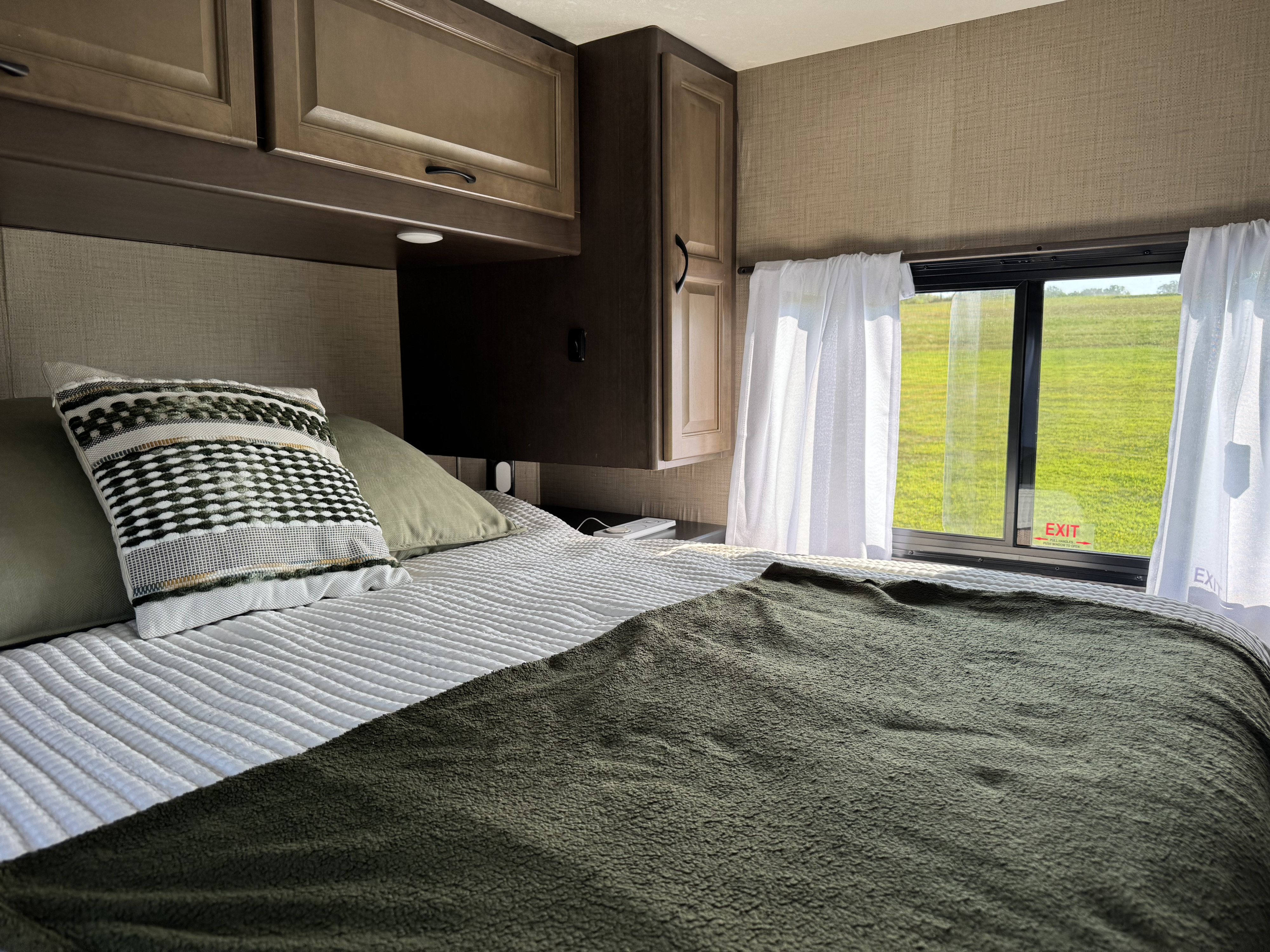 Cozy RV rental bedroom with white bedding, olive green accents, and a clear view of a green field.. Thor Majestic 2019